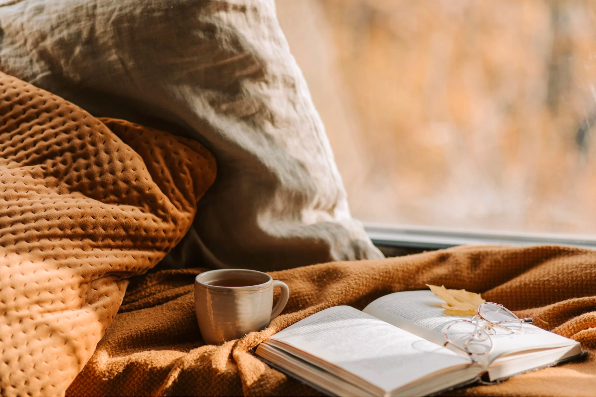 A cup, an open book and a pair of glasses are placed on an orange textured blanket