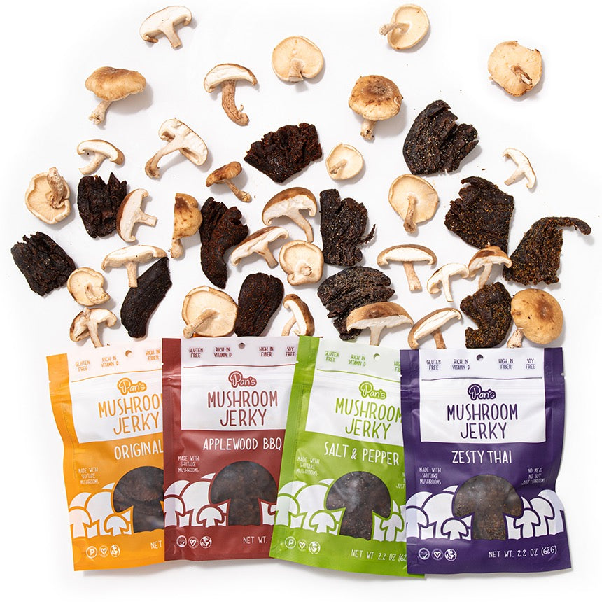 A collection of Pan’s Mushroom Jerky in various flavors against a white background with pieces of raw mushrooms and jerky within the flat lay. 