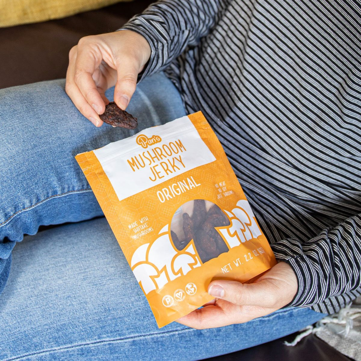 A model’s hands holds a bag of Pan’s Mushroom Jerky against their outfit of jeans and a strippped shirt. 