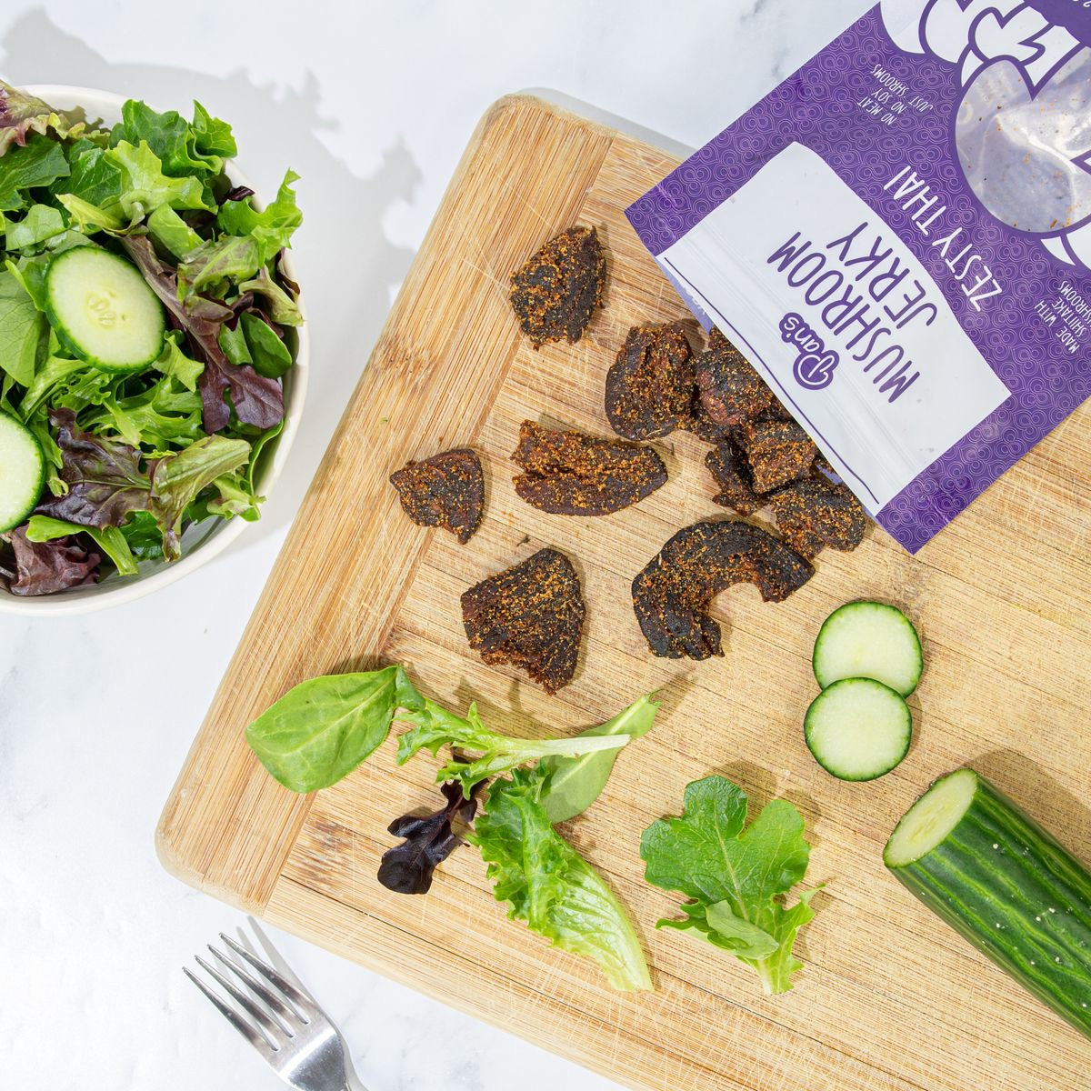 A bag of zesty Pan’s Mushroom Jerky against a cutting board along with some veggies and a fork. 