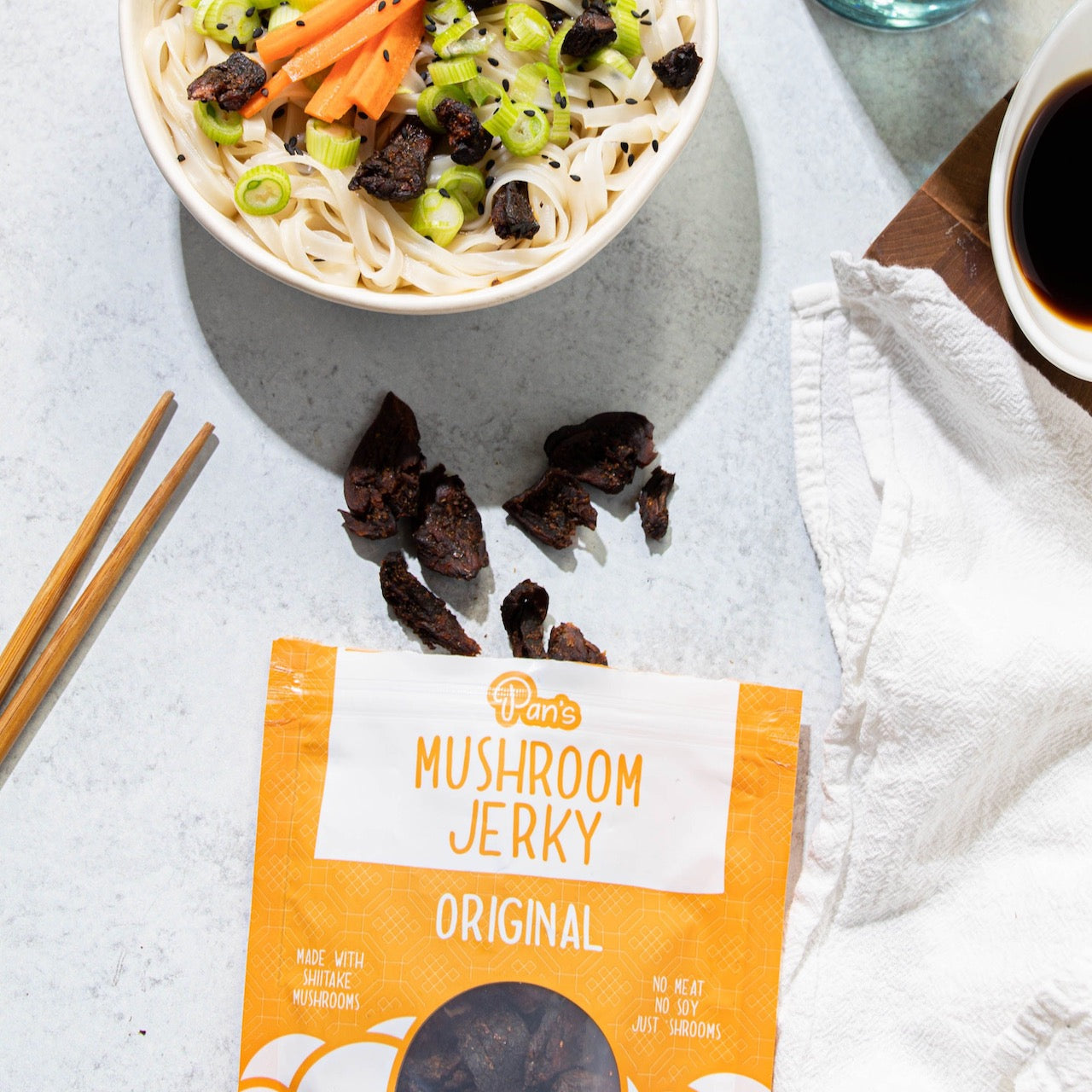 A bag of original flavored Pan’s Mushroom Jerky below a bowl of noodles along with some chopsticks and a container of sauce. 