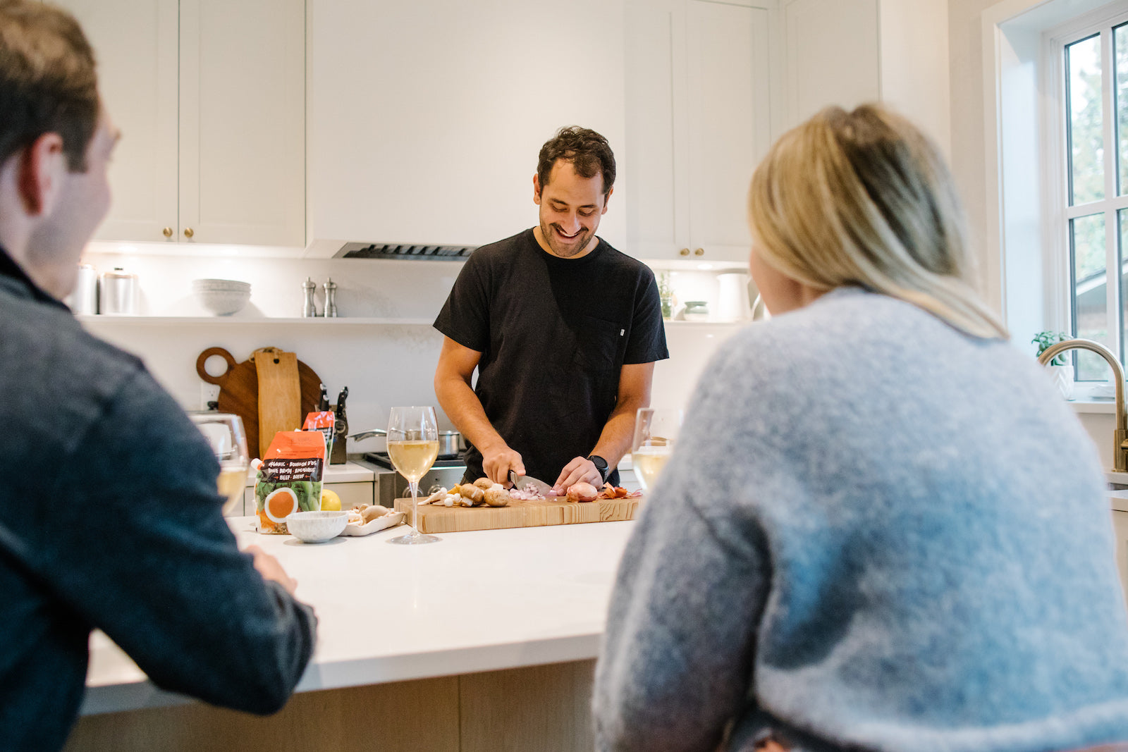 Two guests sit in a kitchen while a male customer of Bluebird Provisions is making a meal in the background. 