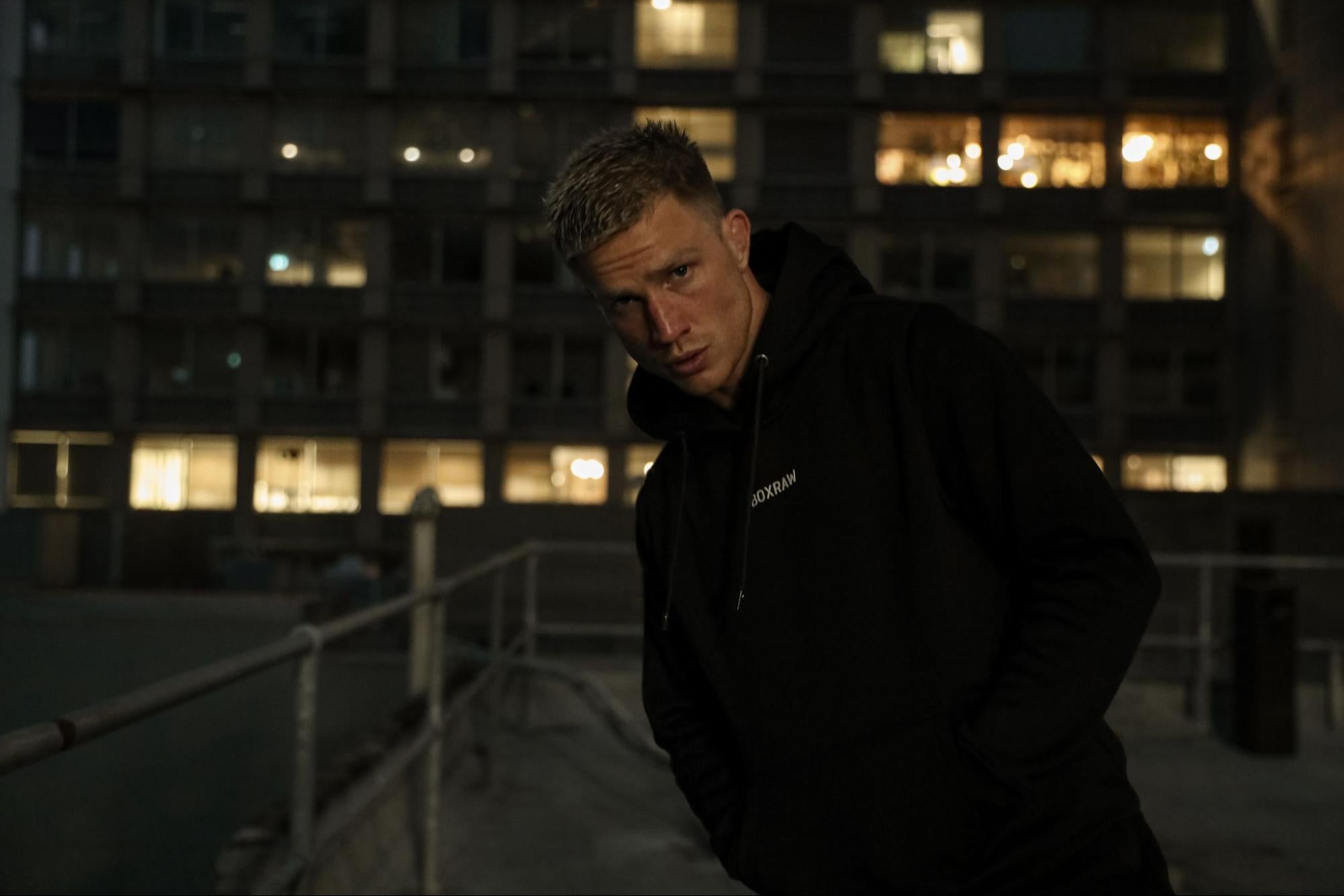 A male model wears a black hoodie by Boxraw backdropped by apartment buildings. 