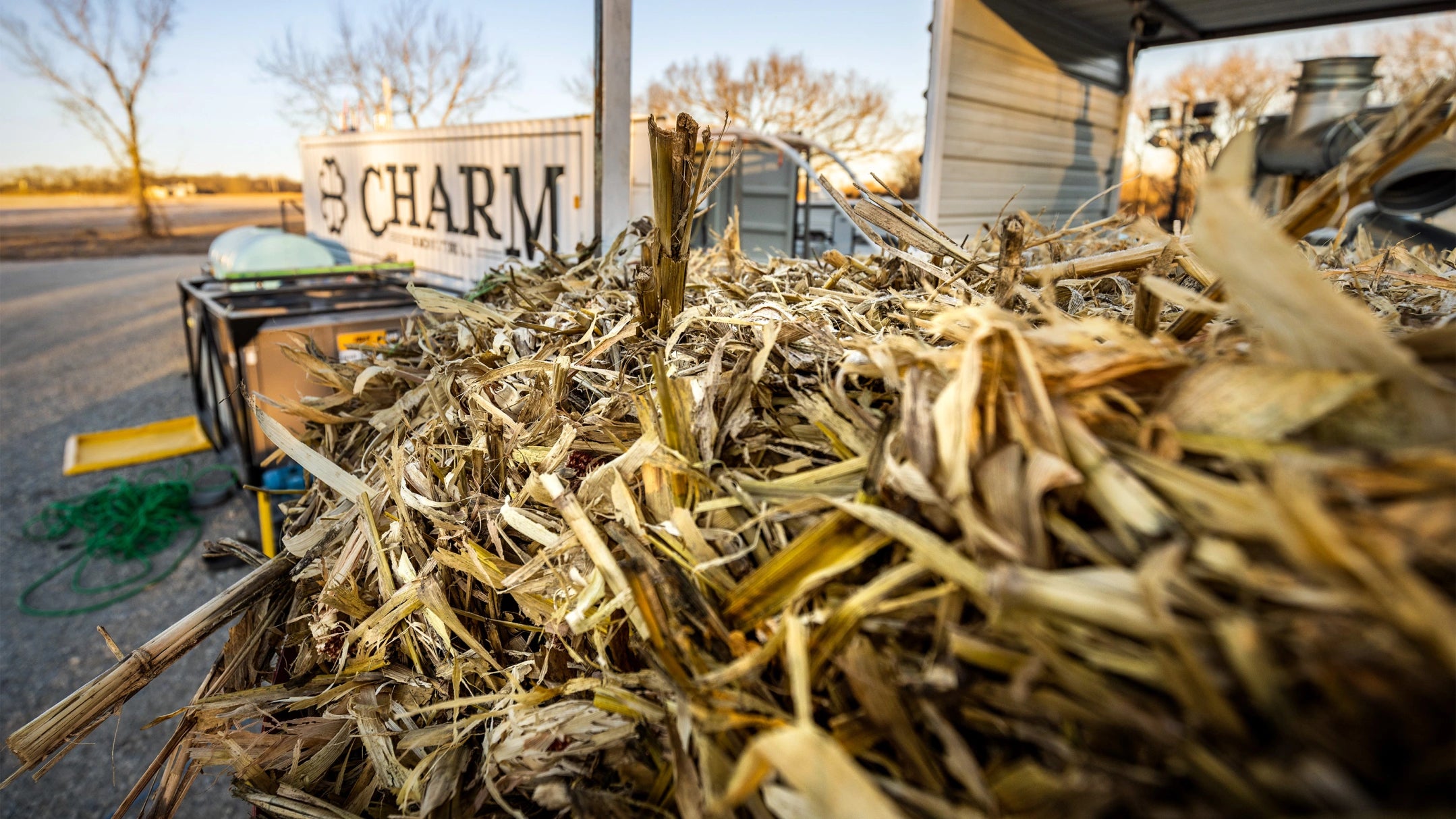 Pile of organic matter including dried stalks and leaves. A Charm logo appears in the background