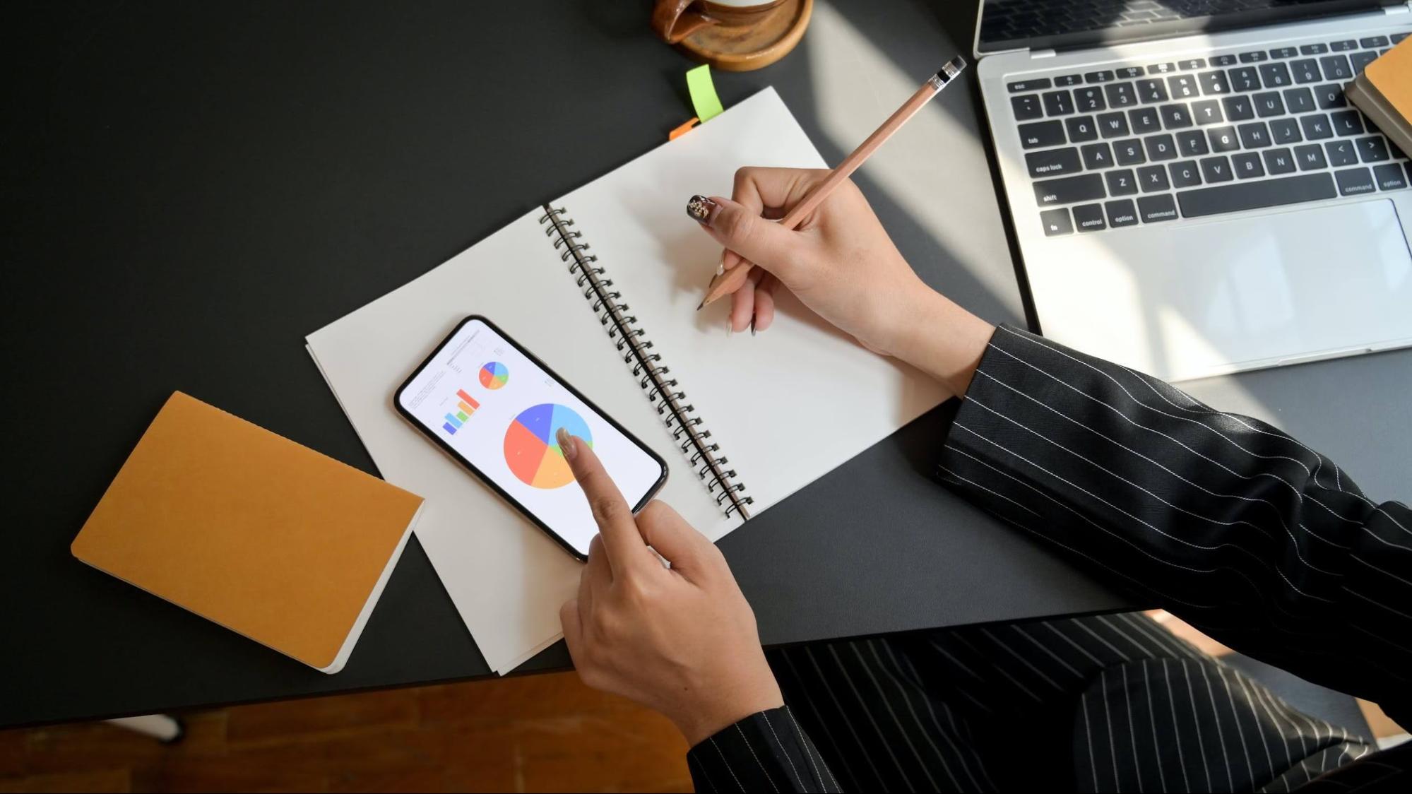 A person sits at a table, writing in a notebook while checking charts on a phone.