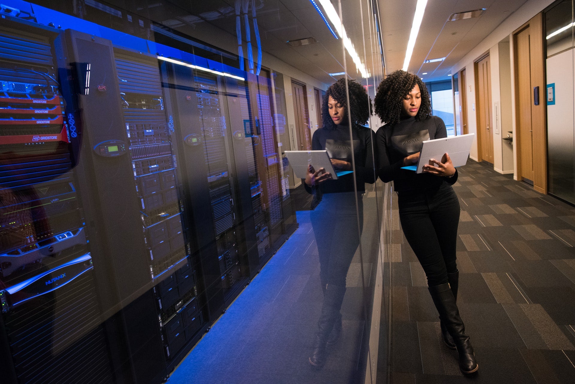 woman in capeted hallway holding laptop standing against glass window of server room