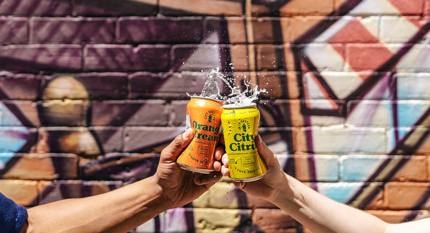 Two people cheers cans of seltzer before a graffitied wall.