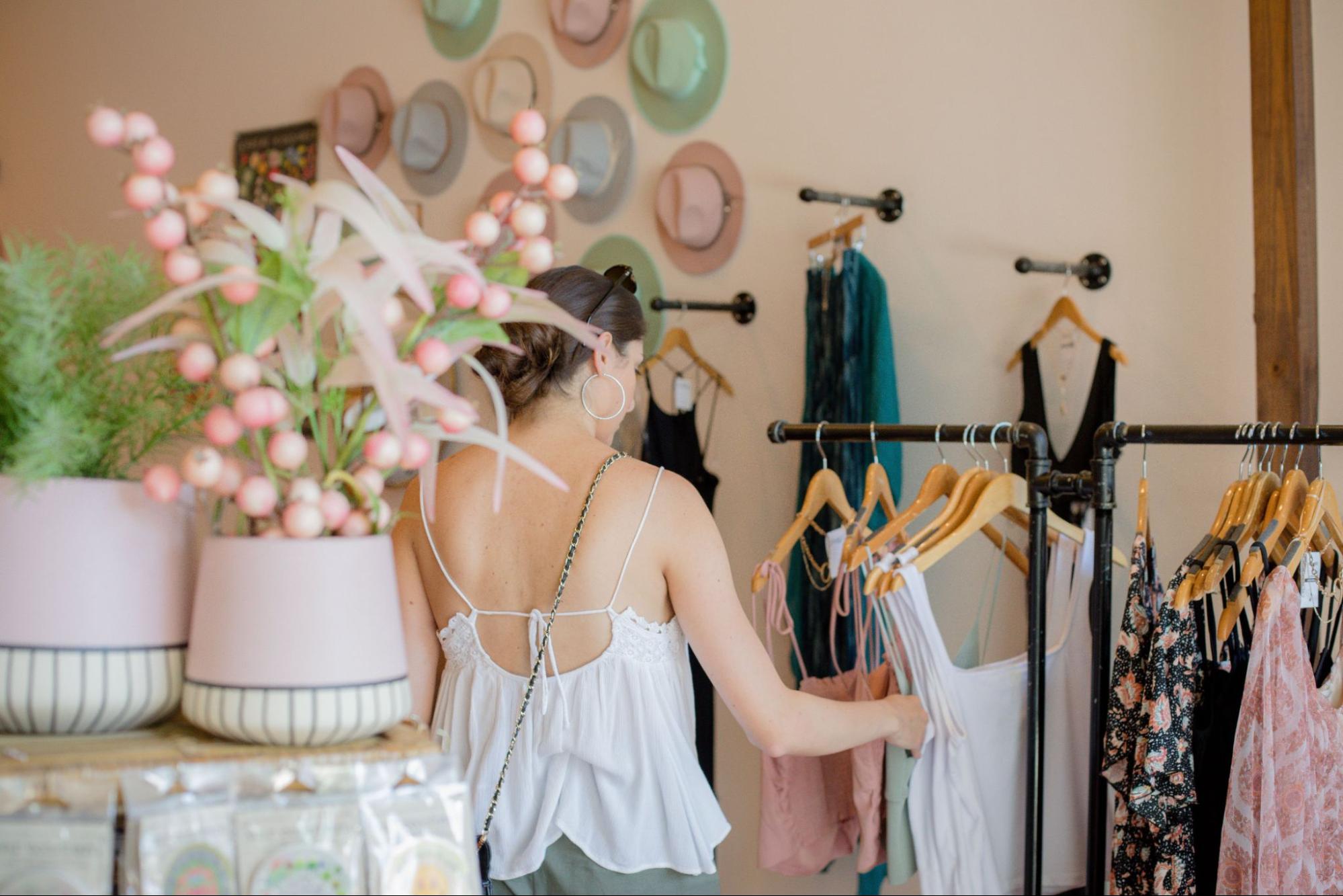 Person in a white top browses racks of clothing in a store. There are two pink pots to the left of the frame with plants in them.