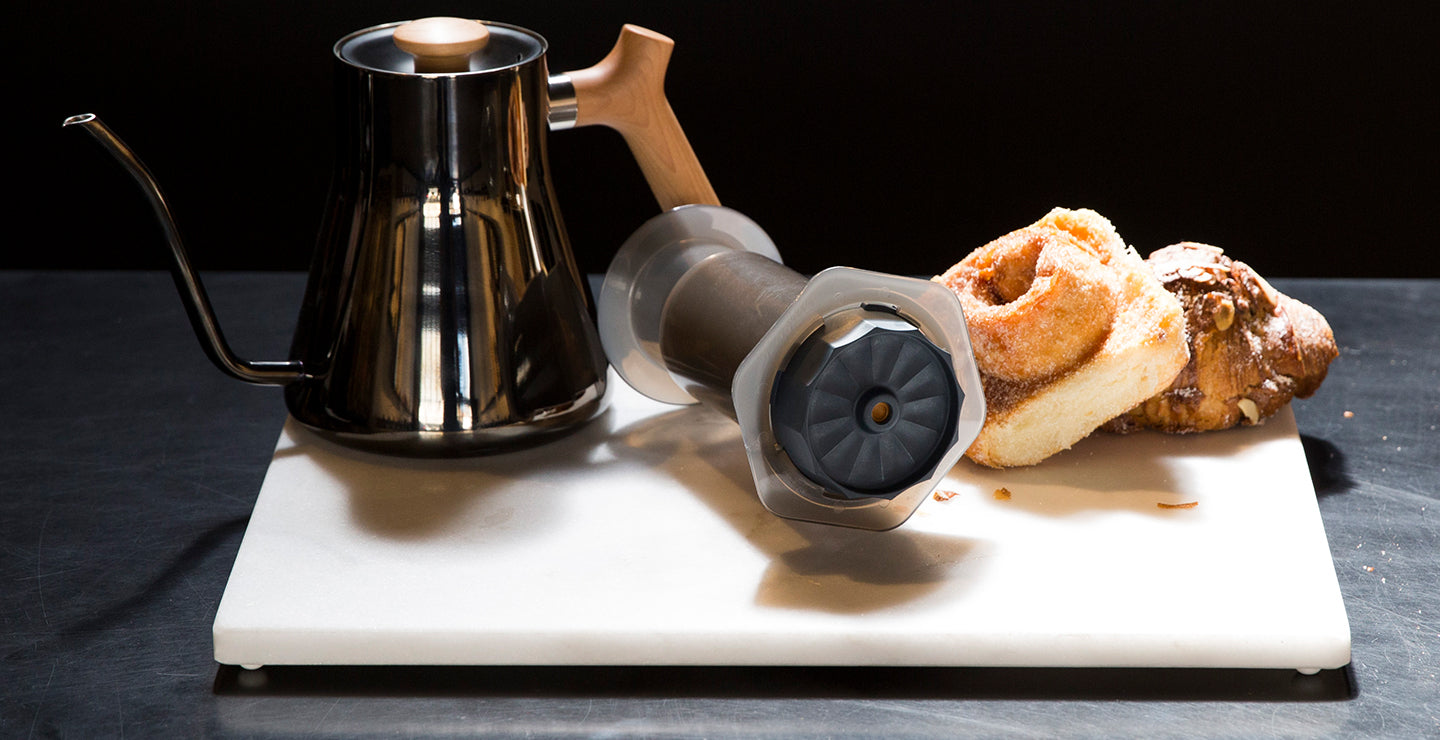 The Prismo attachment for AeroPress laying on a cutting board next to baked goods and a Fellow kettle. 