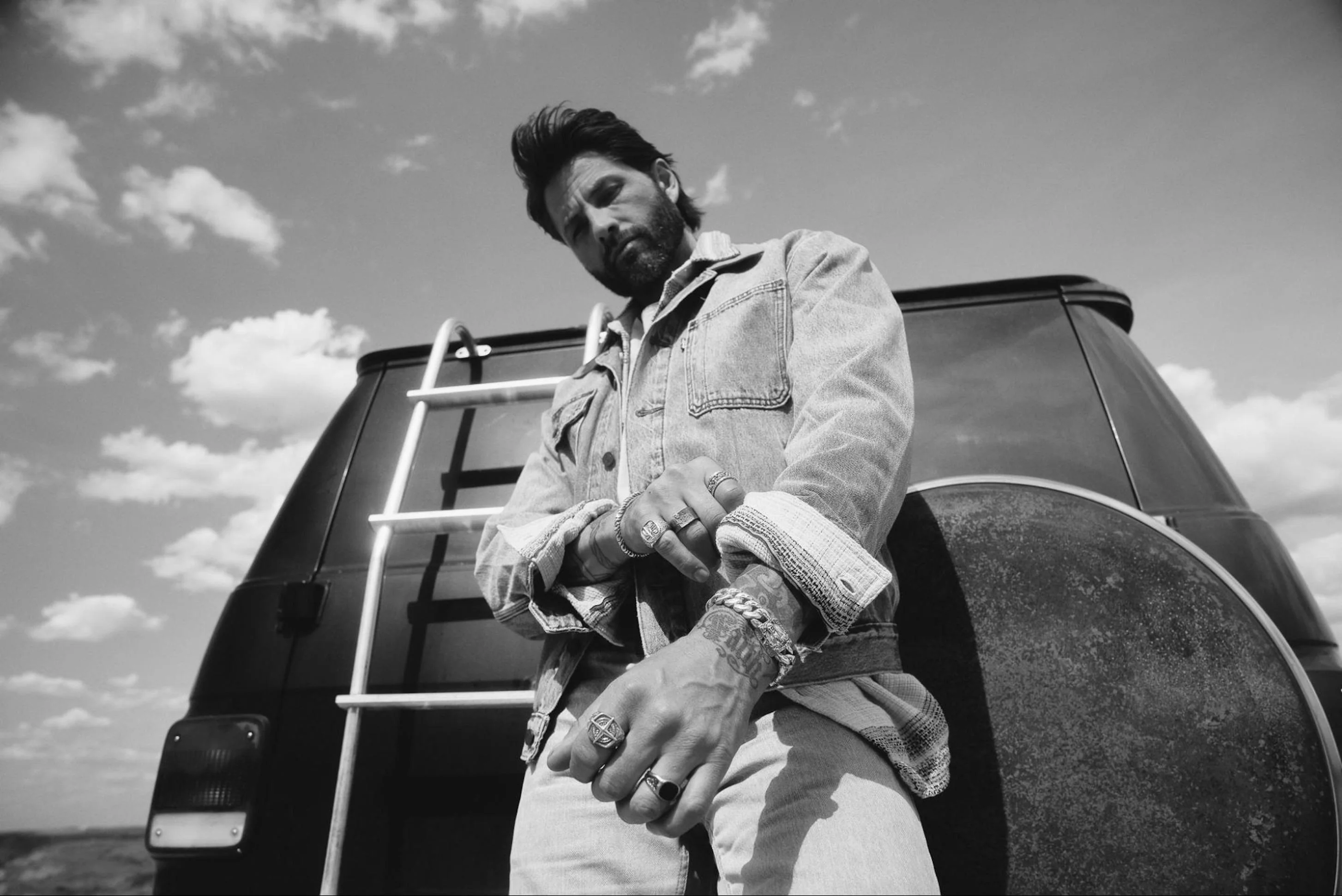 Greyscale photo of a man standing in the dessert behind a caravan rolling up his jacket sleeve to reveal Clocks and Colours rings and bracelets, plus a tattoo.