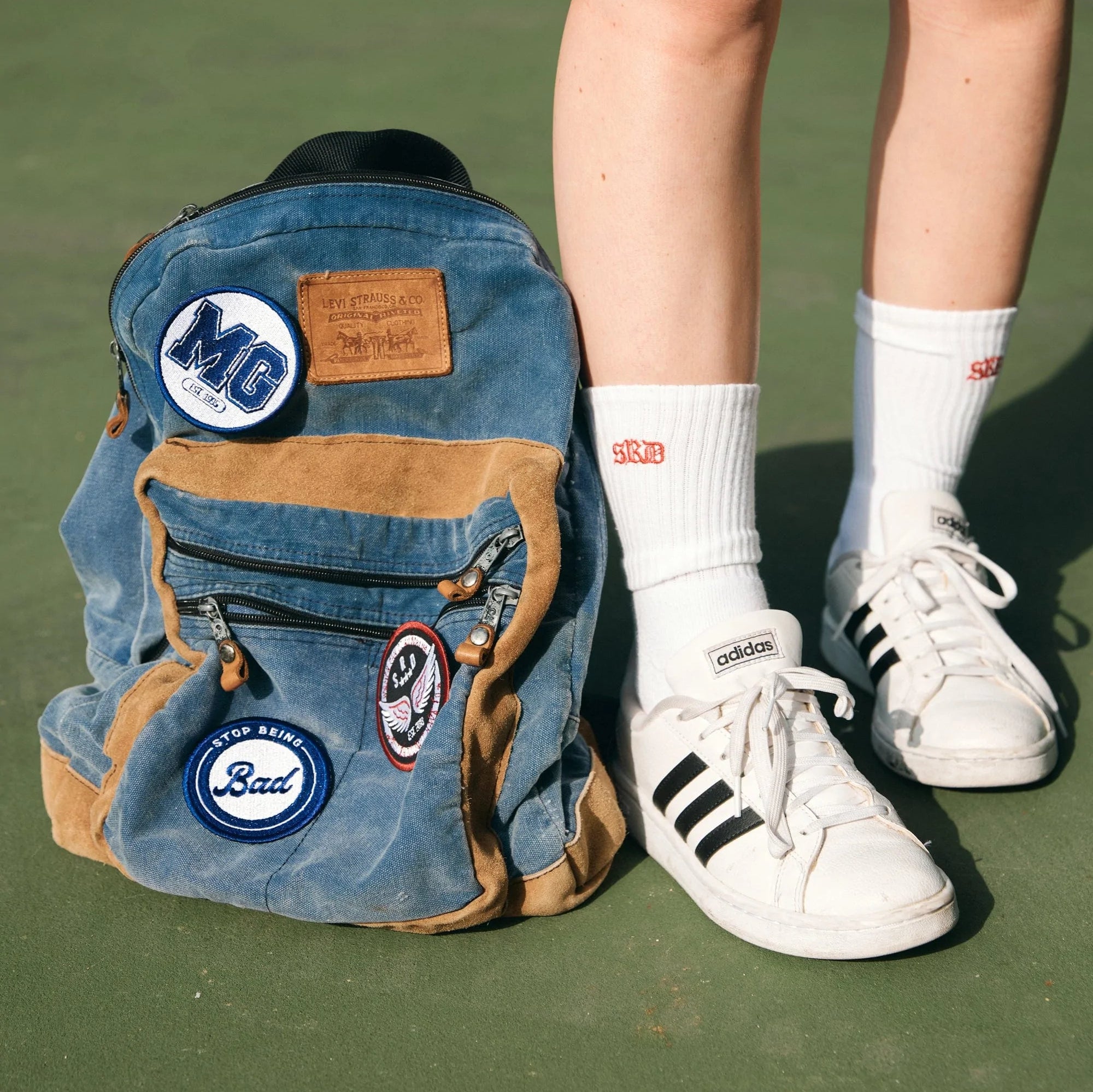 A person stands beside a backpack with patches, and is pictured from the knees down