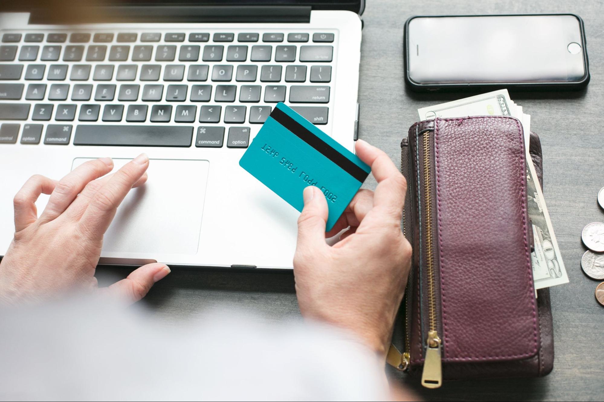 A hand holds a credit card they have pulled from their wallet to pay for something on their laptop.