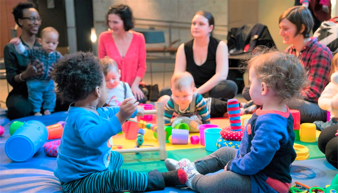 Infants and daycare workers hang out on a play mat with brightly colored toys.