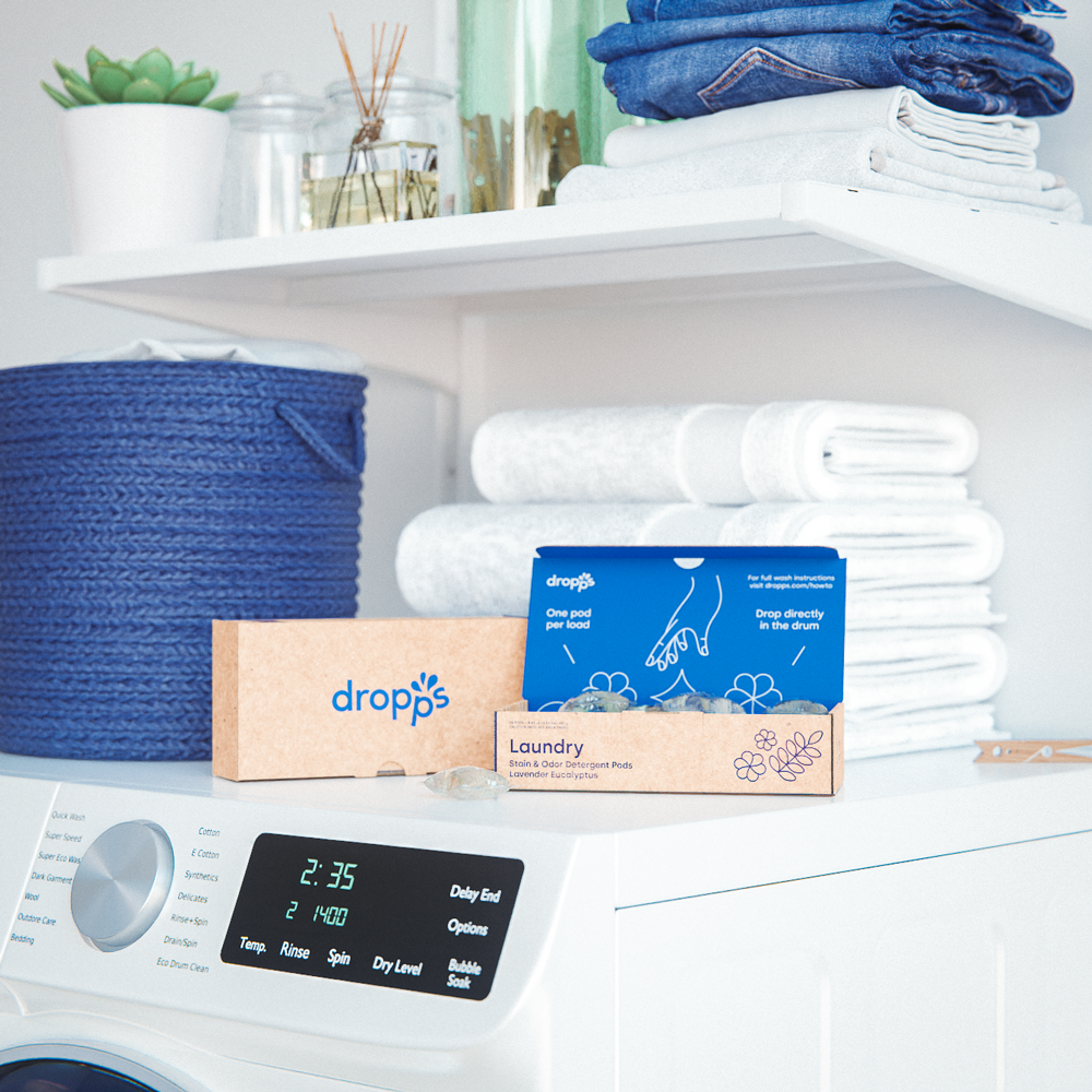 A Dropps package positioned on a laundry machine backdropped by folded towels and a blue basket.