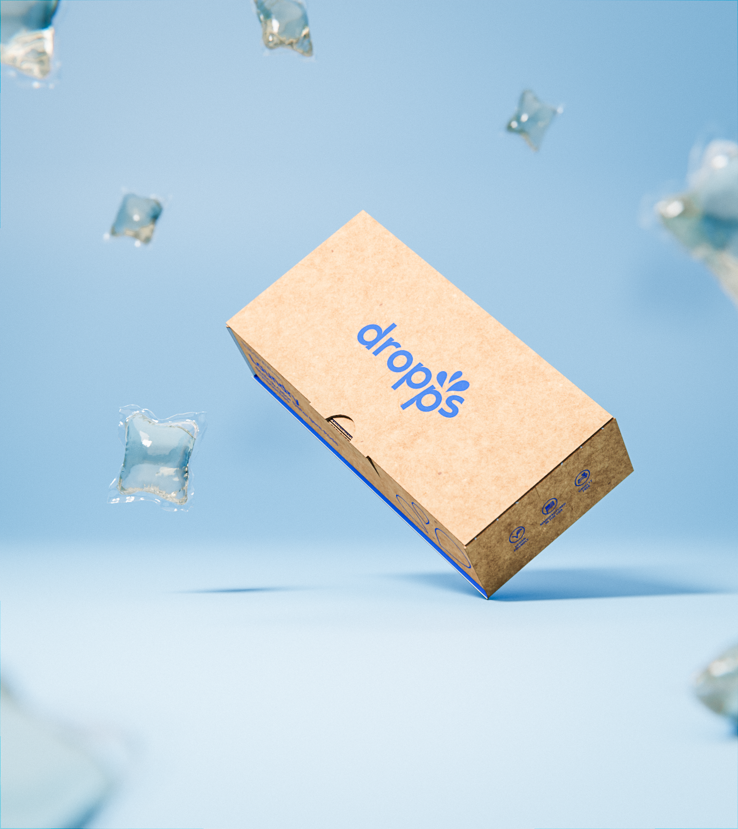 A box of dropps suspended in the air against a light blue background, surrounded by clear plastic pods that are also suspended. 