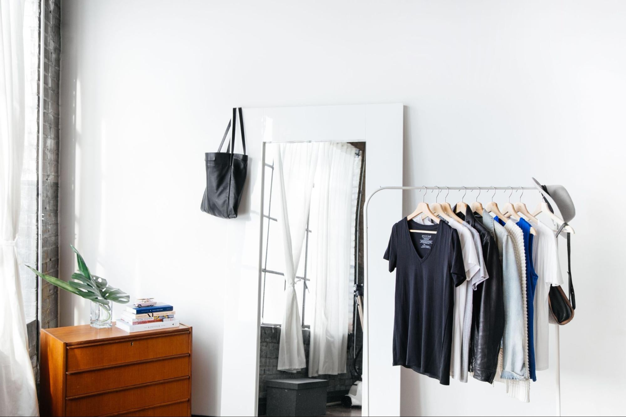 A rack of clothing by Enrcicled in the same room as a full-length mirror and dresser. 