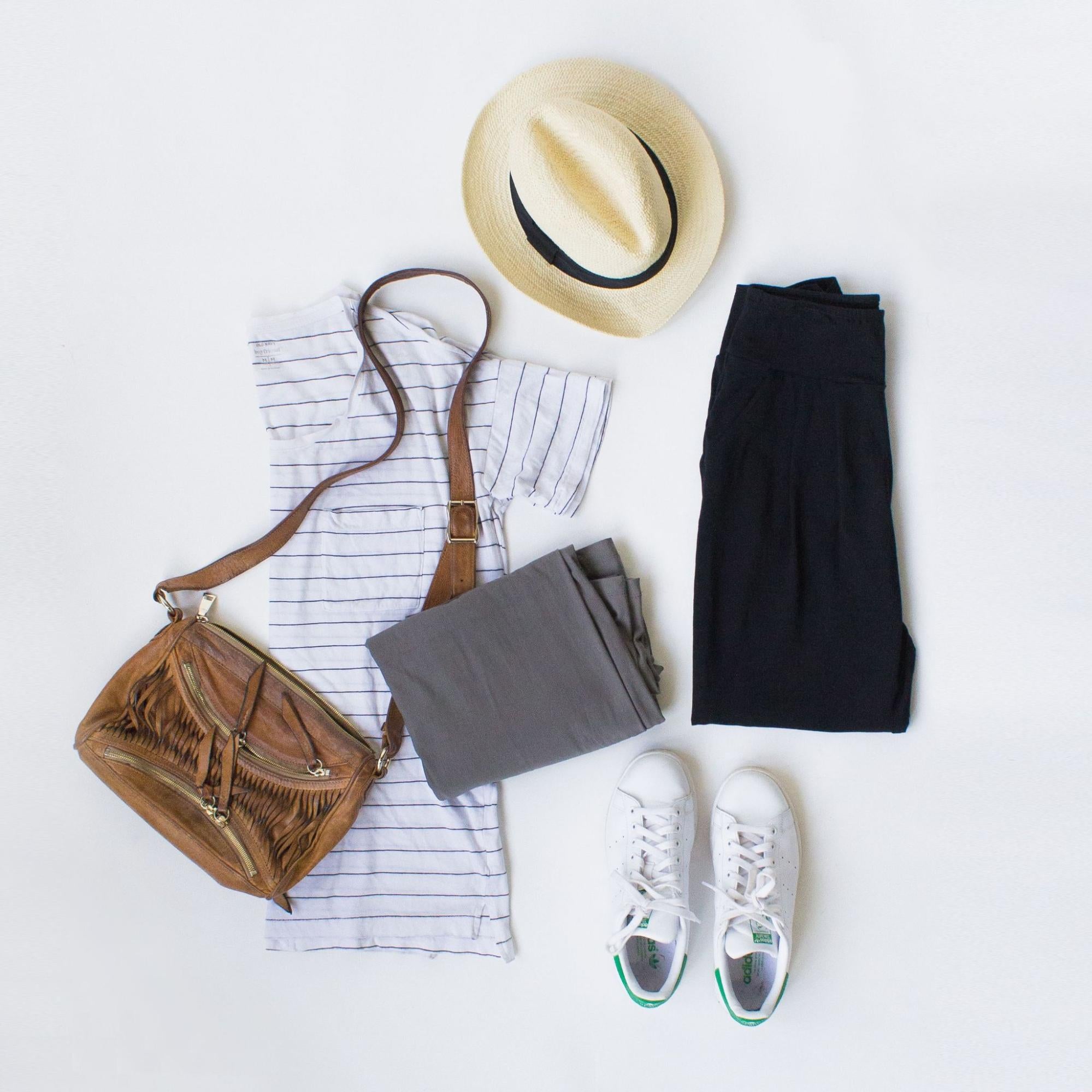 A flat lay of a shirt, two pairs of pants, shoes, a crossbody purse and a sun hat.