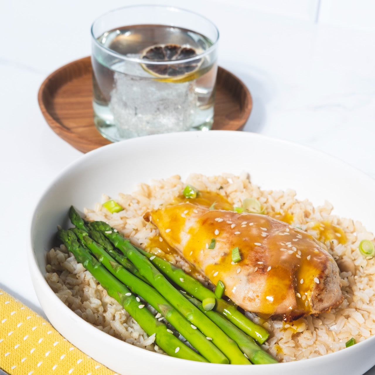 A plate with chicken, asparagus, and brown rice backdropped by a glass filled with soda water and a dried slice of red grapefruit.