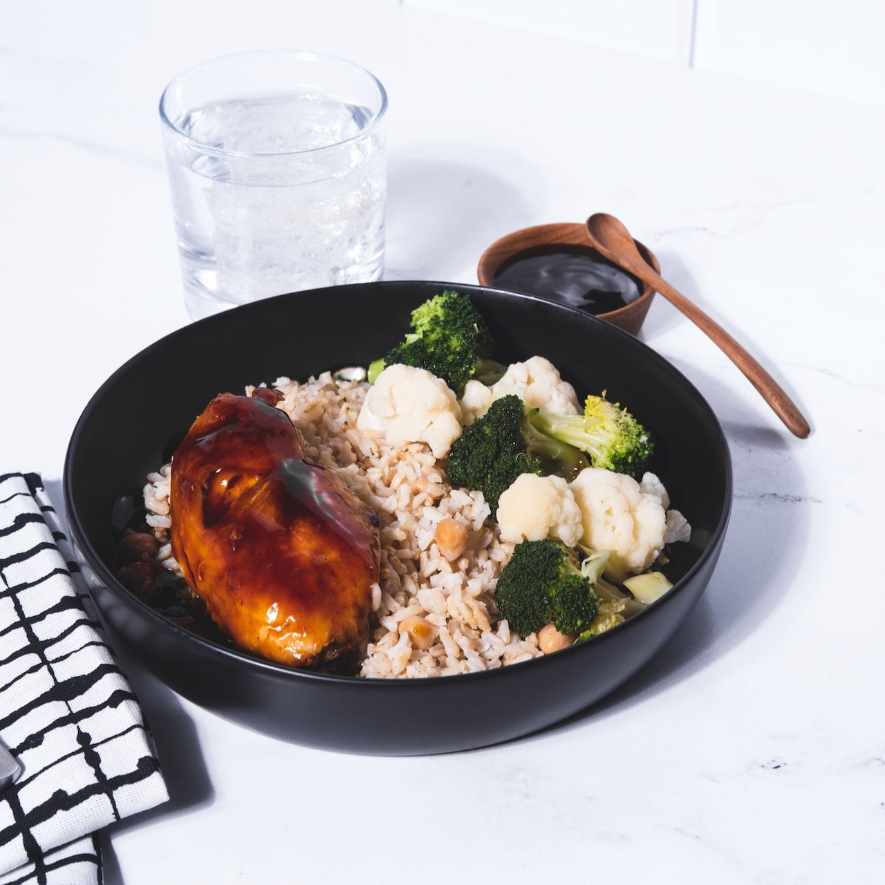 A bowl of broccoli, cauliflower, chicken, and brown rice backdropped by a glass of water and a small ramekin of sauce and a spoon. 