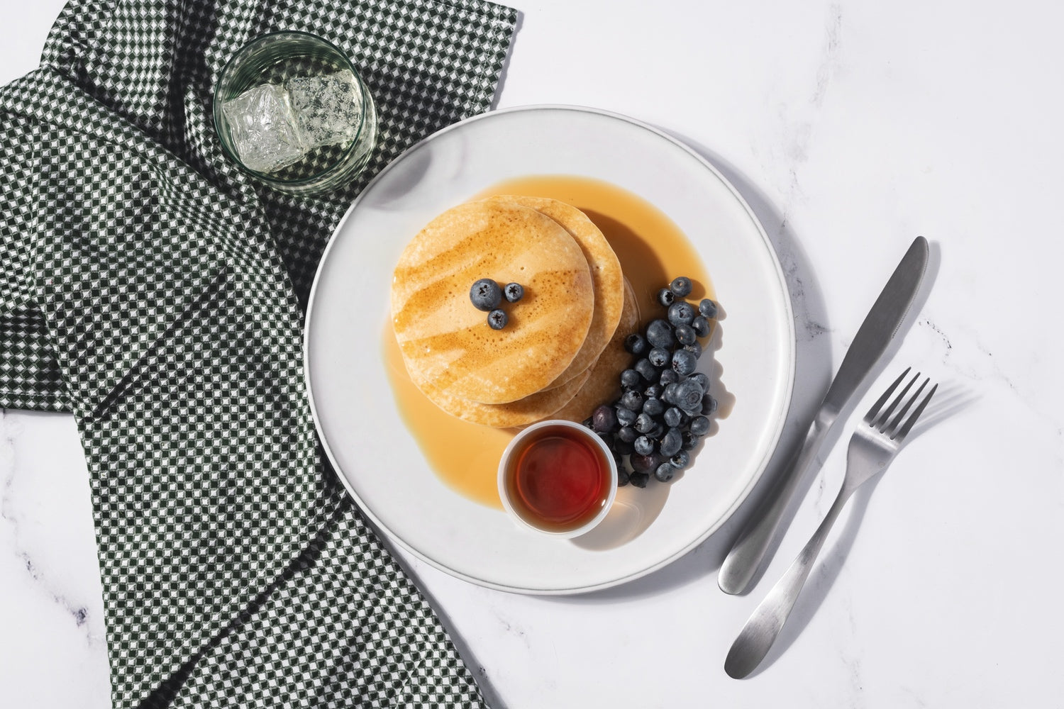 A stack of pancakes on a plate with blueberries and a ramekin of syrup, along with utensils, a napkin, and a glass of water. 