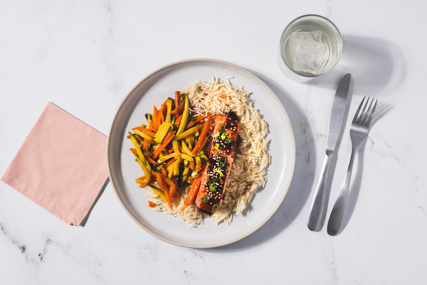 A plate of carrots, zucchini, with salmon and long grain rice backdropped by utensils and a drink in a glass.