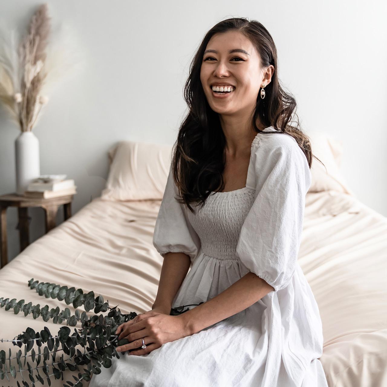 Founder of Eucalypso, Elle Liu, sitting on a bed in a white dress holding branches of eucalyptus.