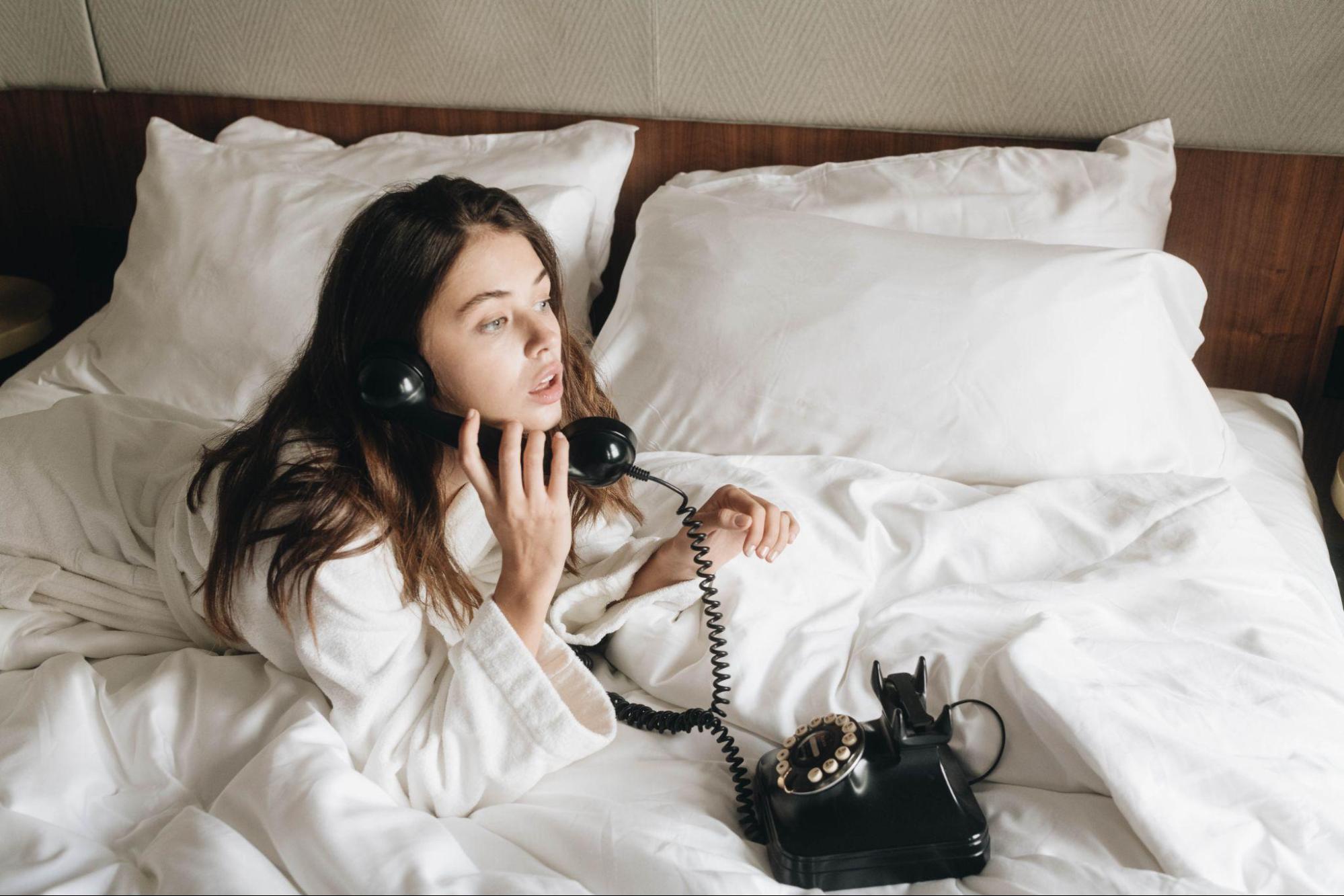 A model in a white bathrobe talking on a rotary phone in a bed with white bedding.