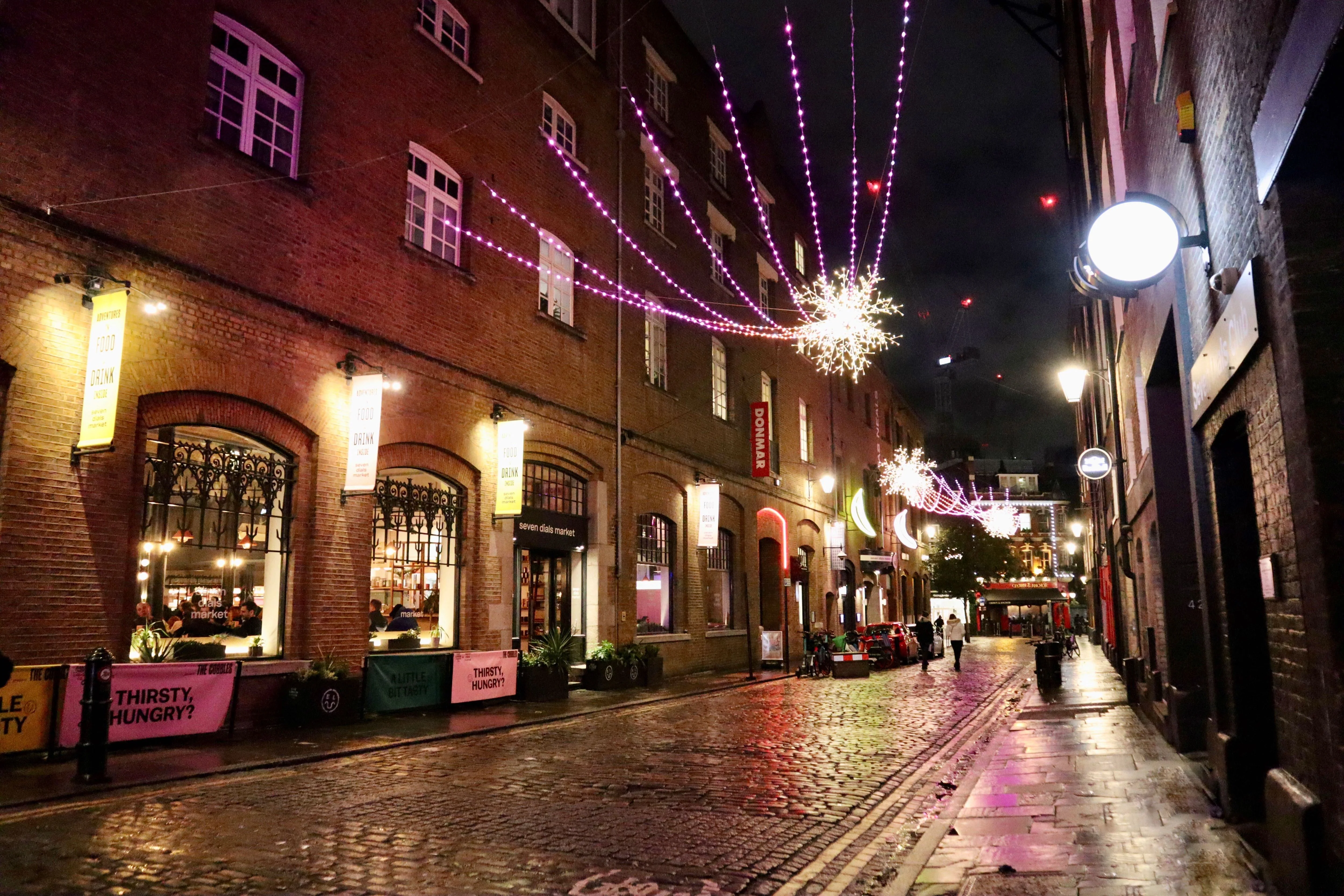 The street view outside of Seven Dials Market during a December evening with holiday lights decorating the building. 