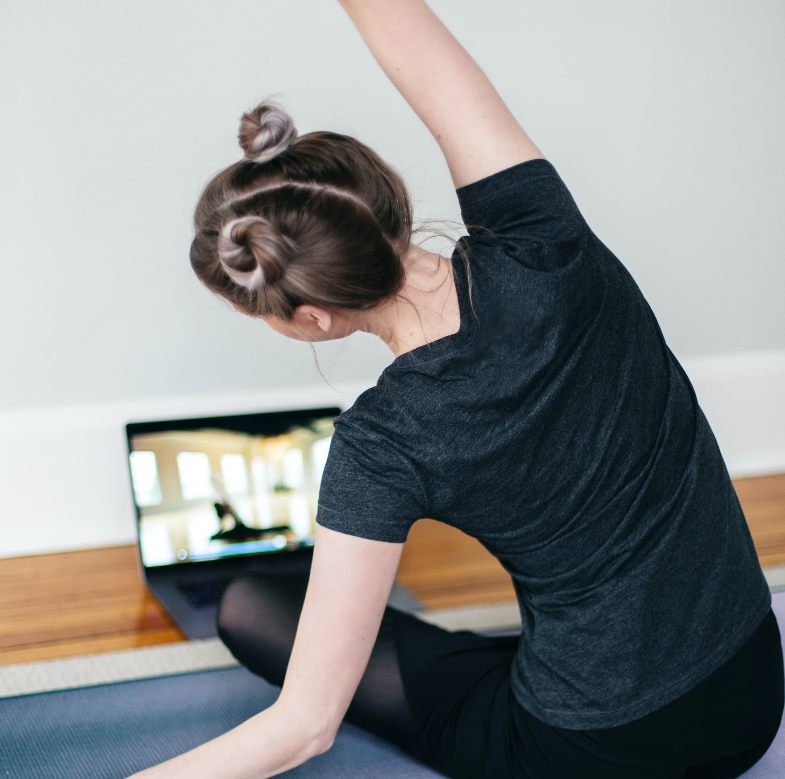 A person sits on a yoga mat watching a livestream yoga class on a laptop