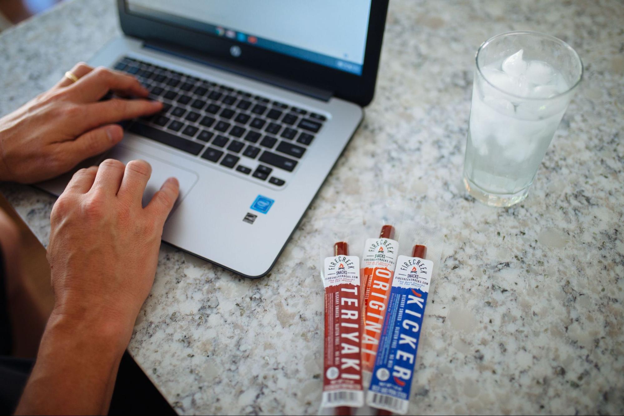 Three jerky from FireCreek Snacks on a marble countertop backdropped by a pair of hands typing on a laptop.