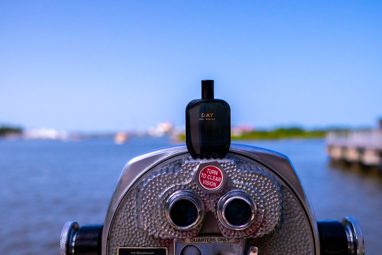 A lookout point backdropped by a body of water and a bottle of Fragrance One perfume rests on top of a tower viewer.