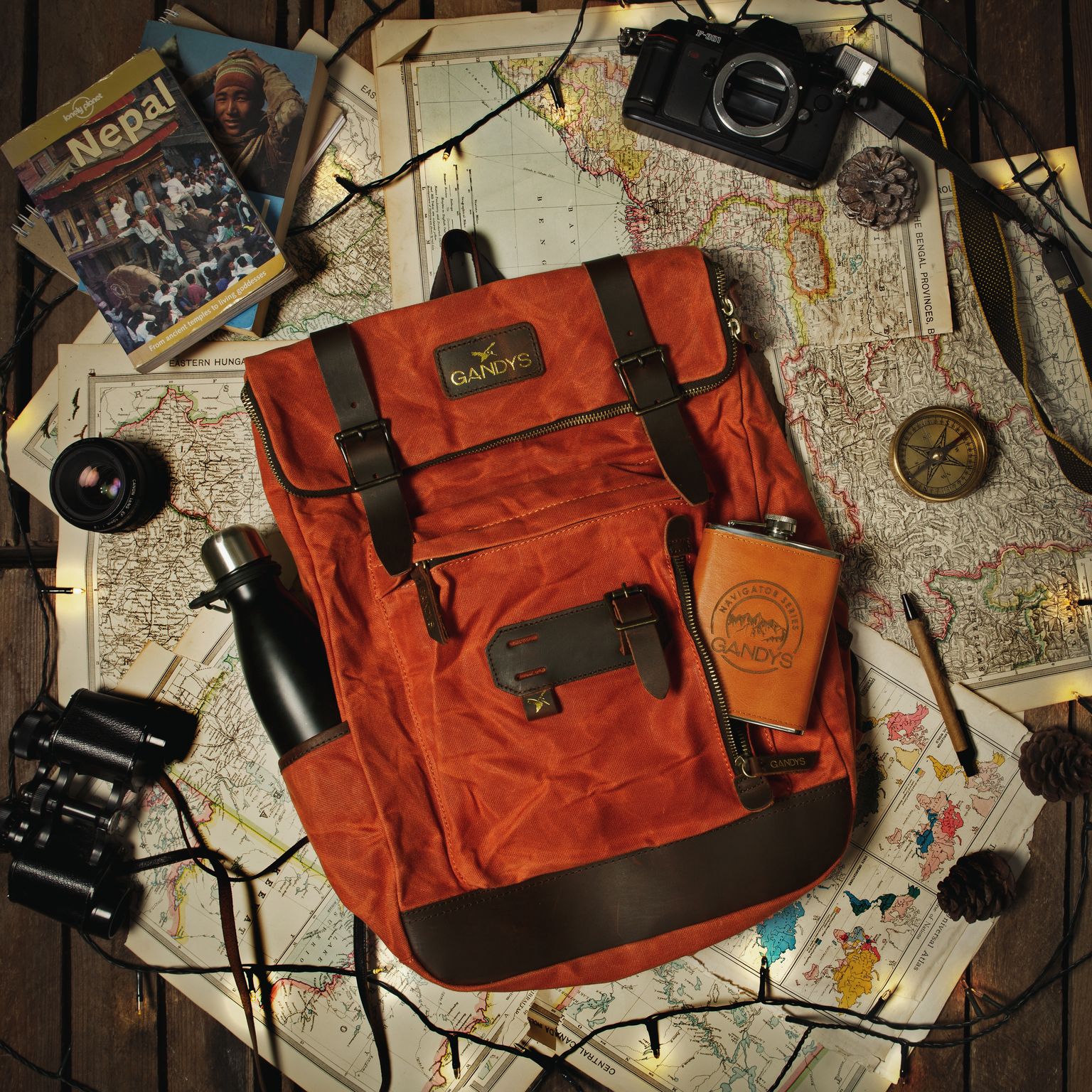 An orange Gandys backpack along with a flask is backdropped by maps and a compass in a flatlay. 