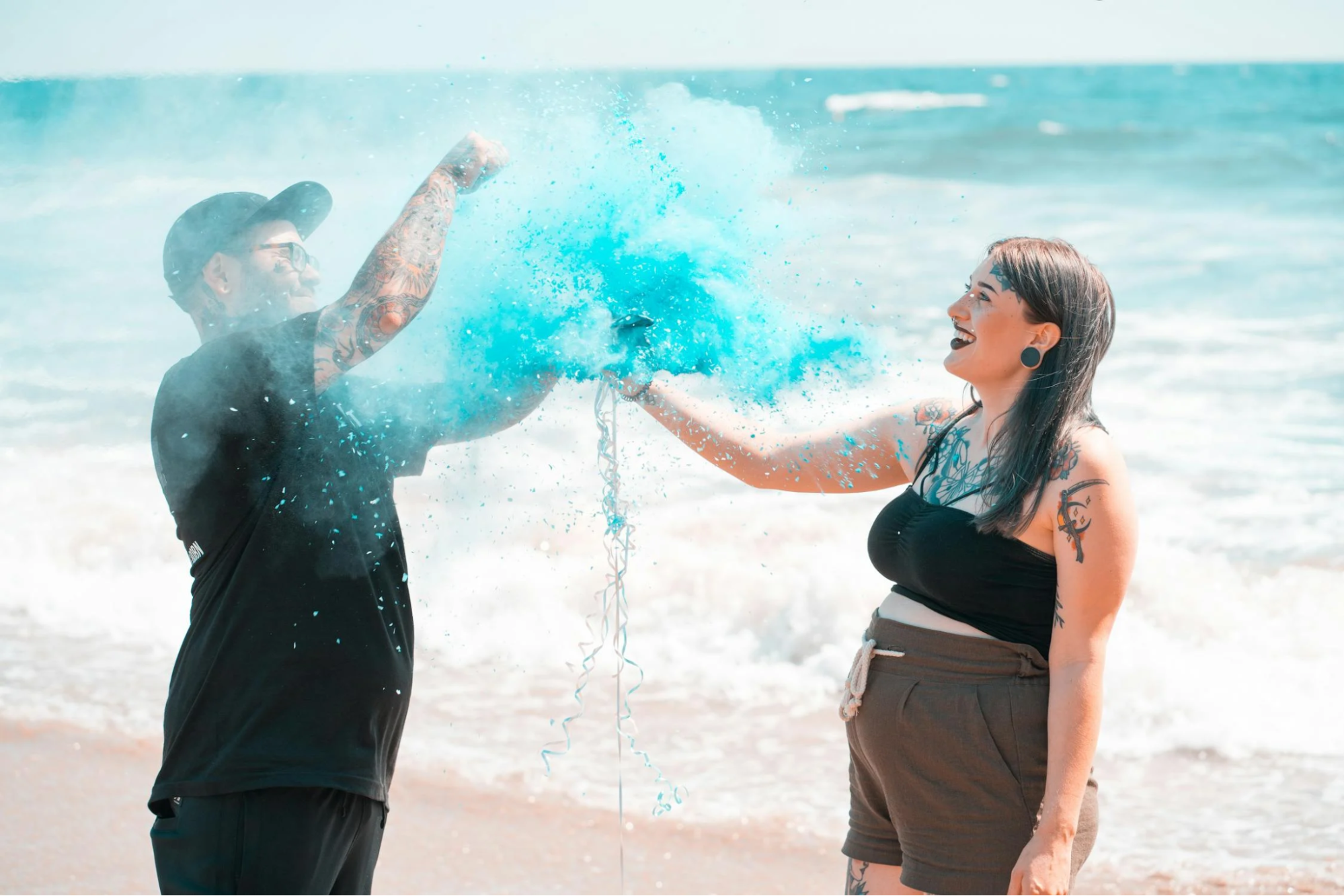 Two expectant parents on a beach pop a gender reveal balloon to release blue glitter.