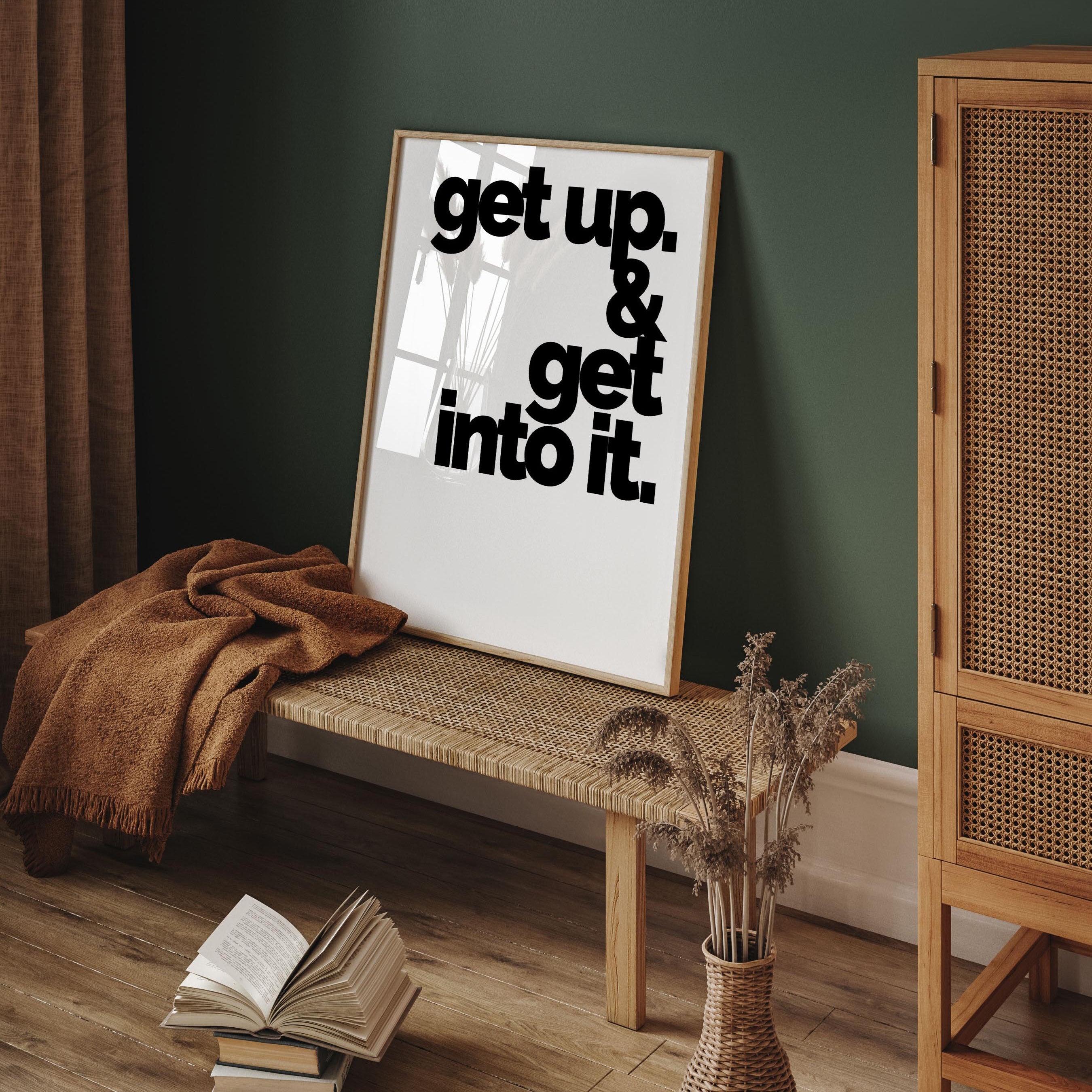 A large framed poster rests on a jute bench in a green room. It reads "get up and get into it"