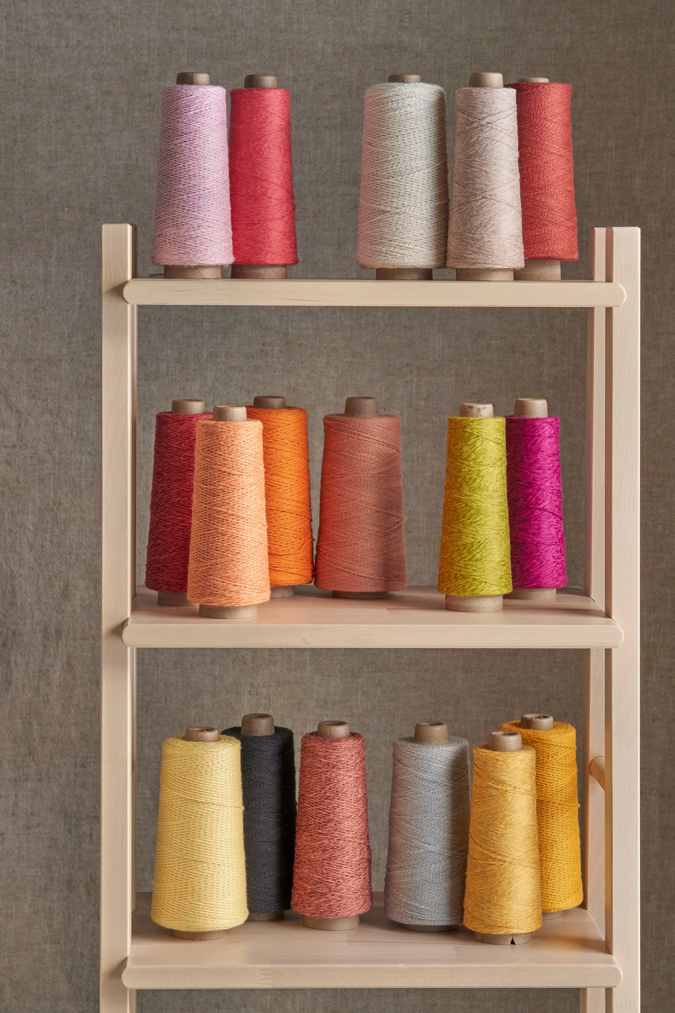 An array of warm toned rolls of threads set on a wooden shelf against a gray background.