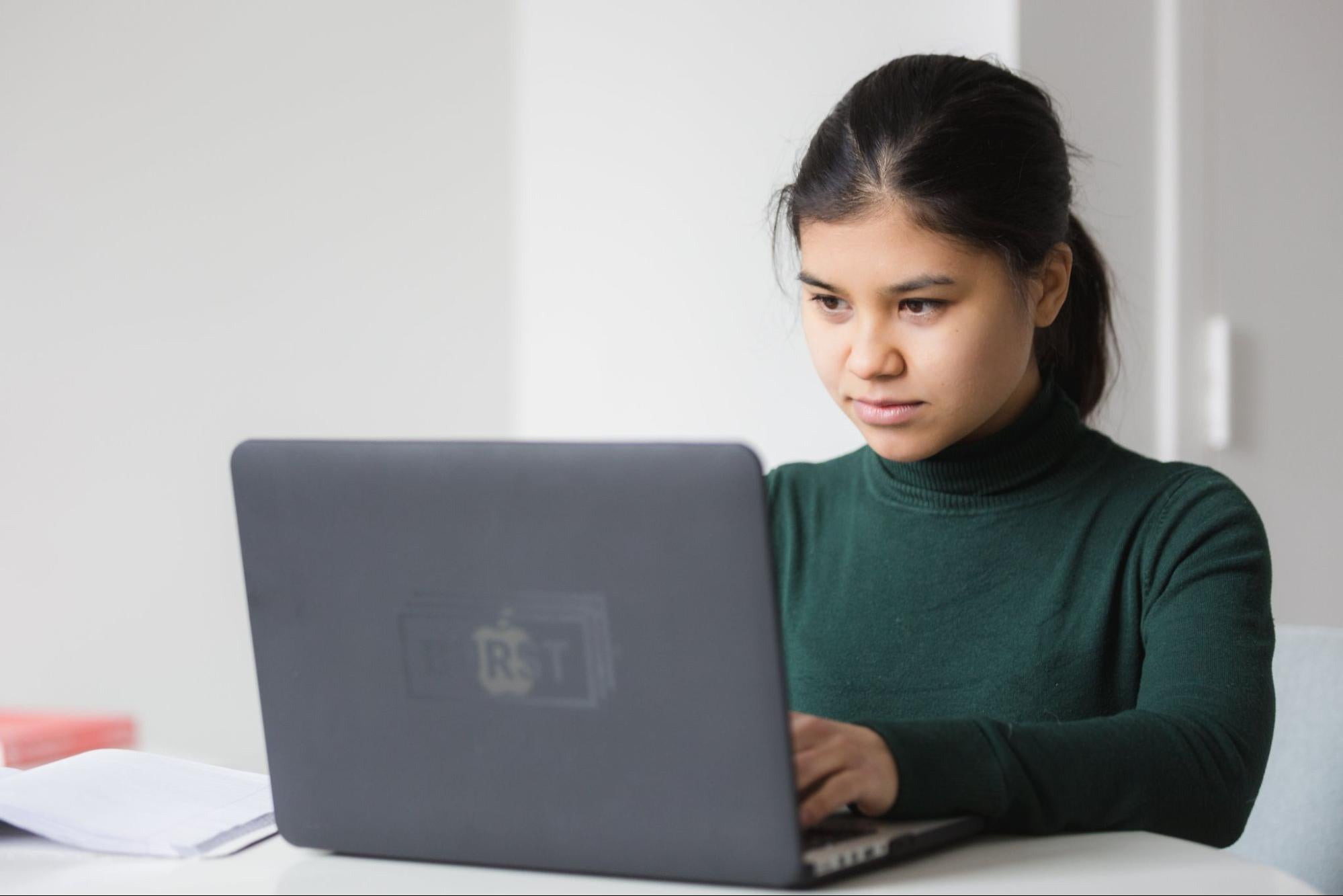 A woman works at her gray laptop on a white table.