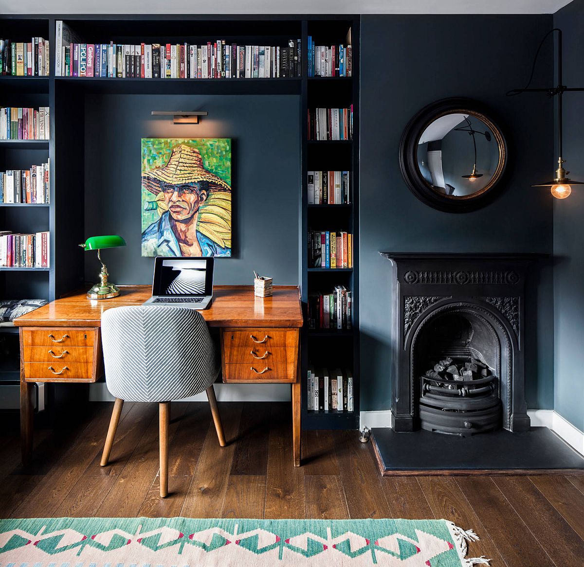 A home office with dark blue walls, vertical bookshelves, bold art and a vintage desk