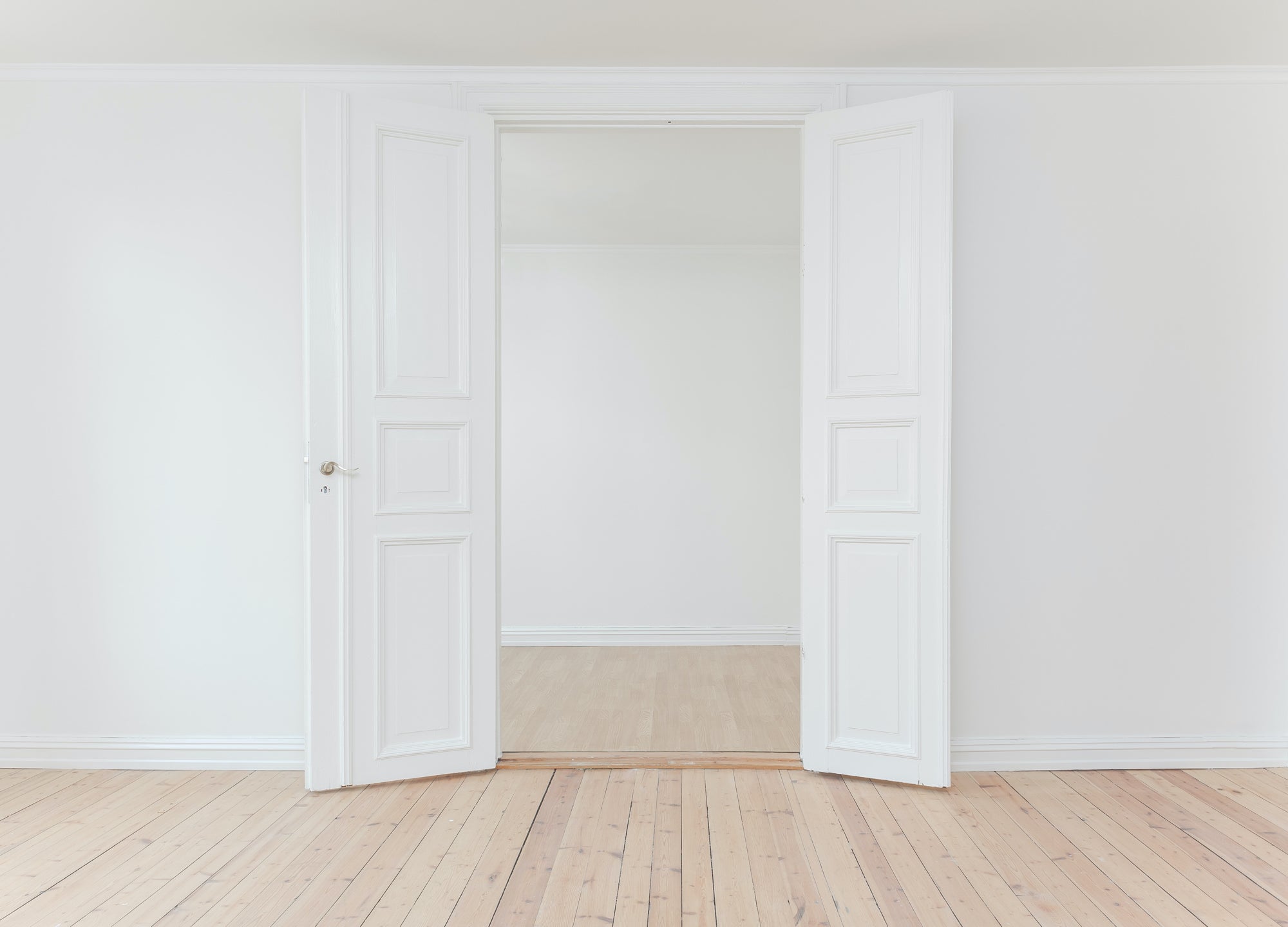 An empty room with wood florrs and white walls looks through a doorway into a similar empty room