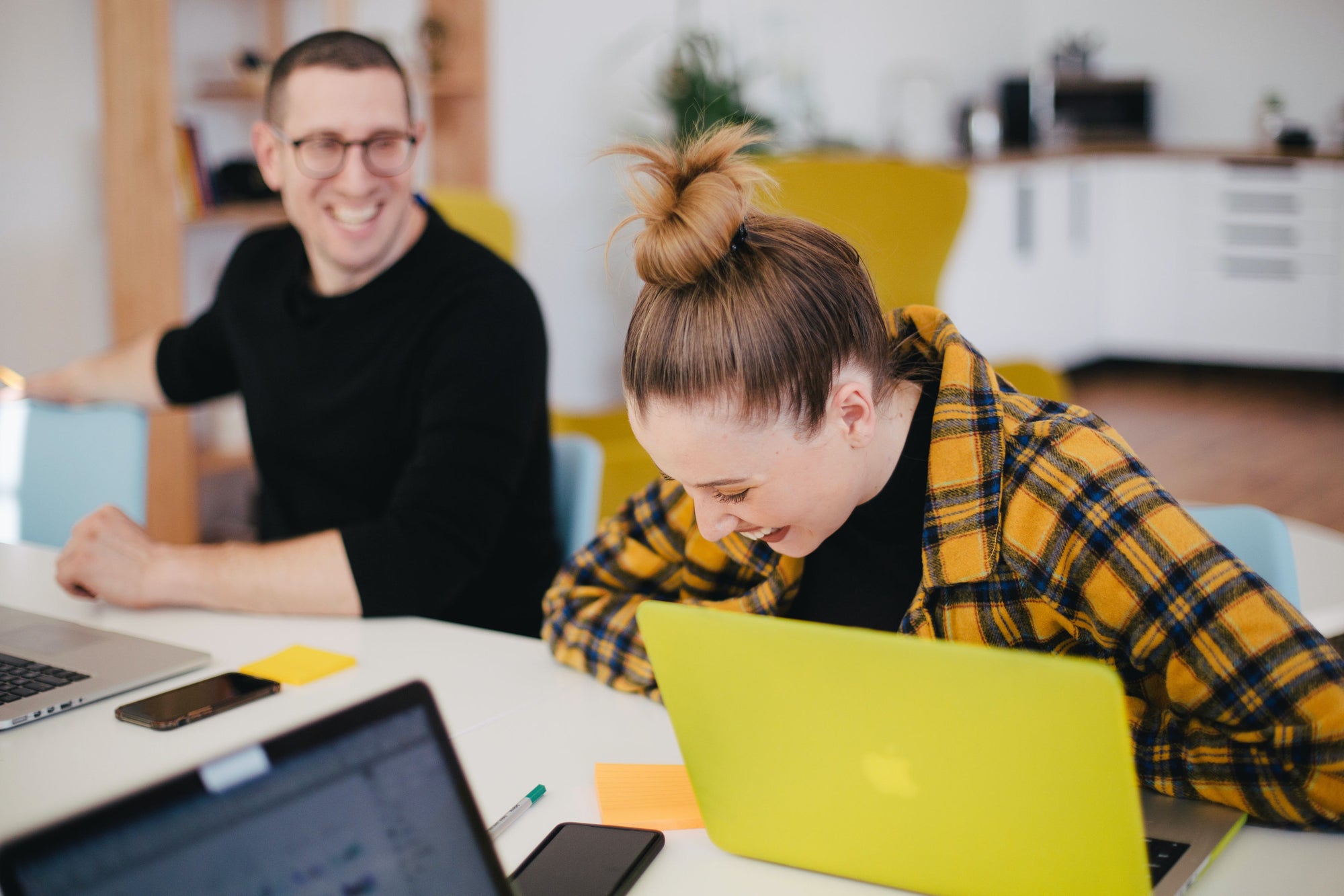 Two people laugh while sitting at a desk with laptops