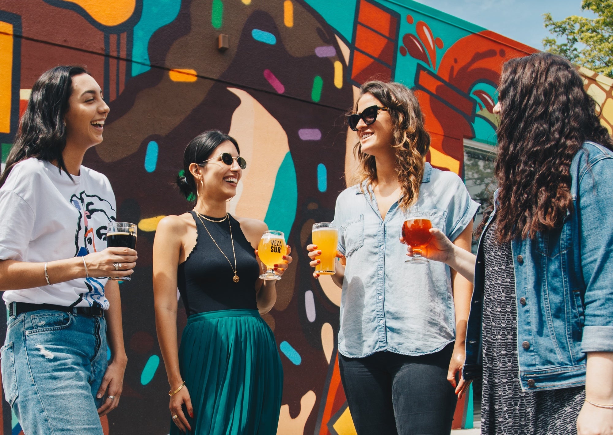 Four people stand holding drinks and socializing in front of an art wall