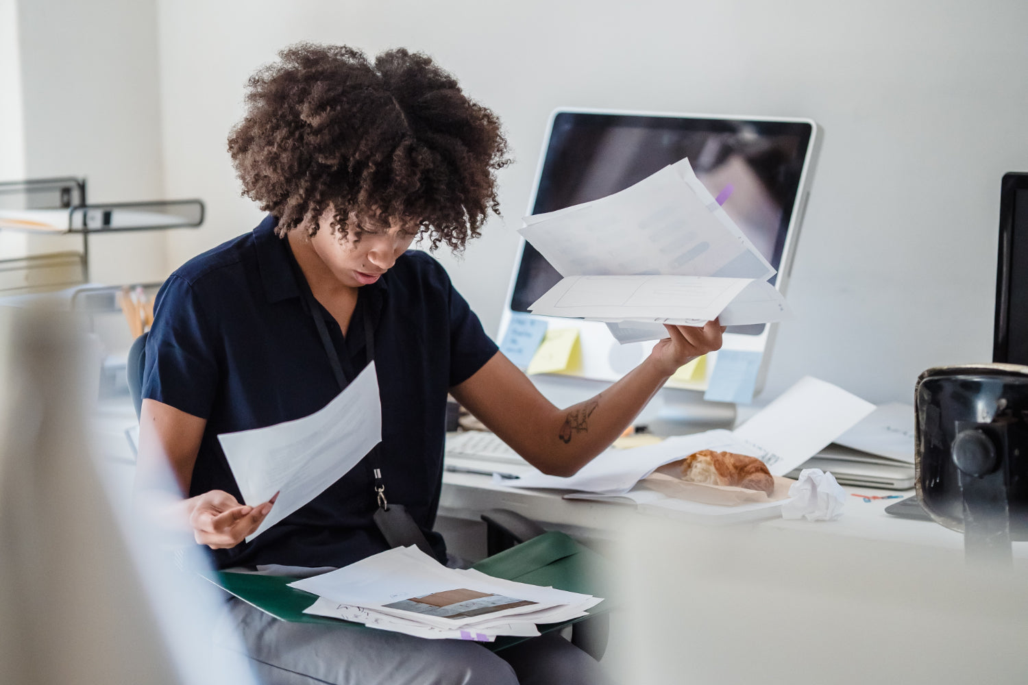 A woman sits at a desk sorting through a stack of papers on her lap