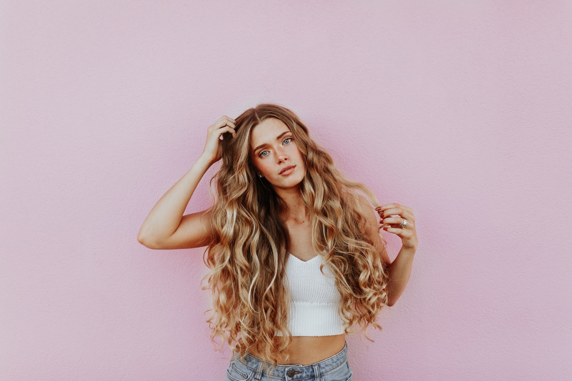 A person with very long curly hair stands against a pink wall