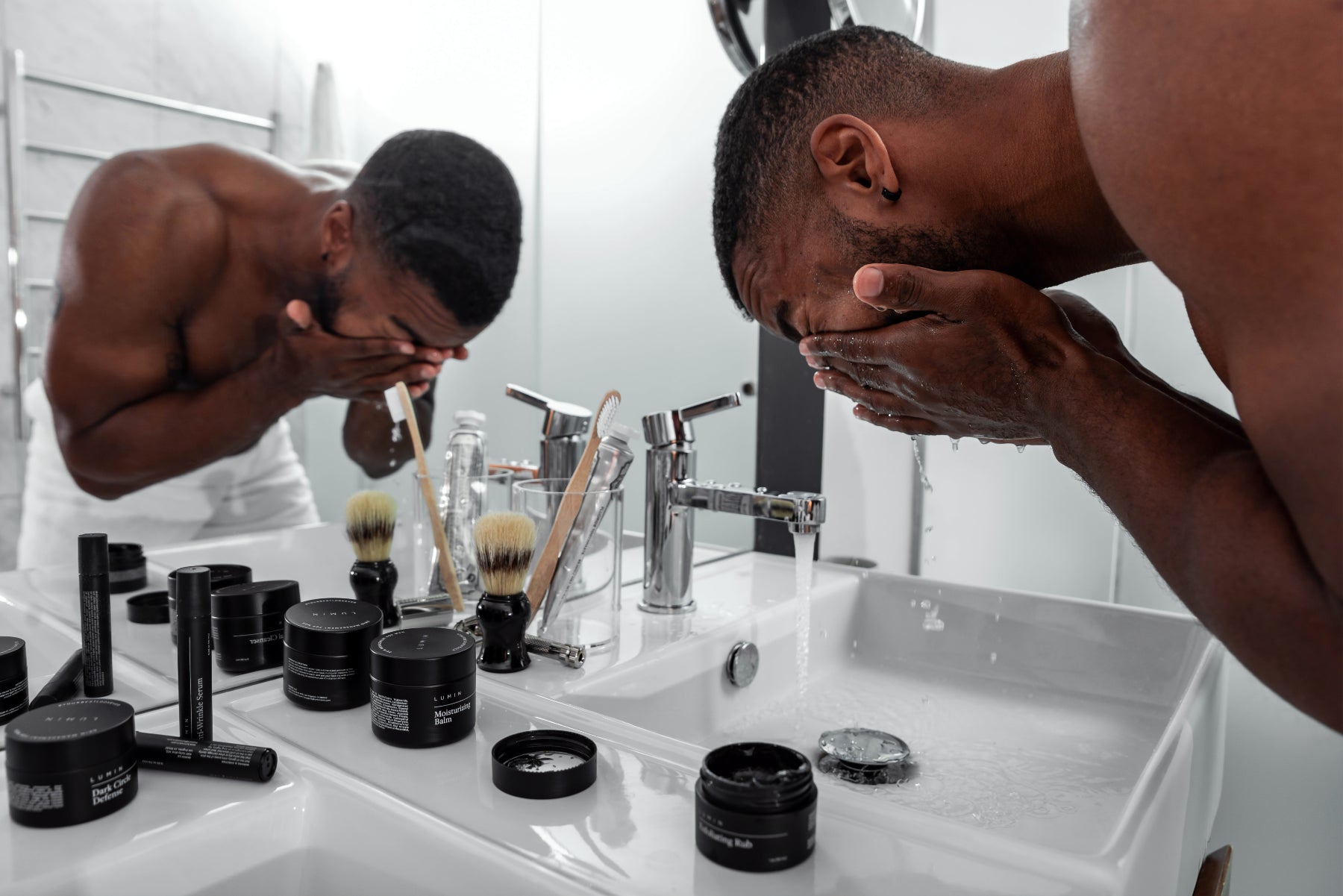 A person washes their face at the sink. Skin products sit on the counter