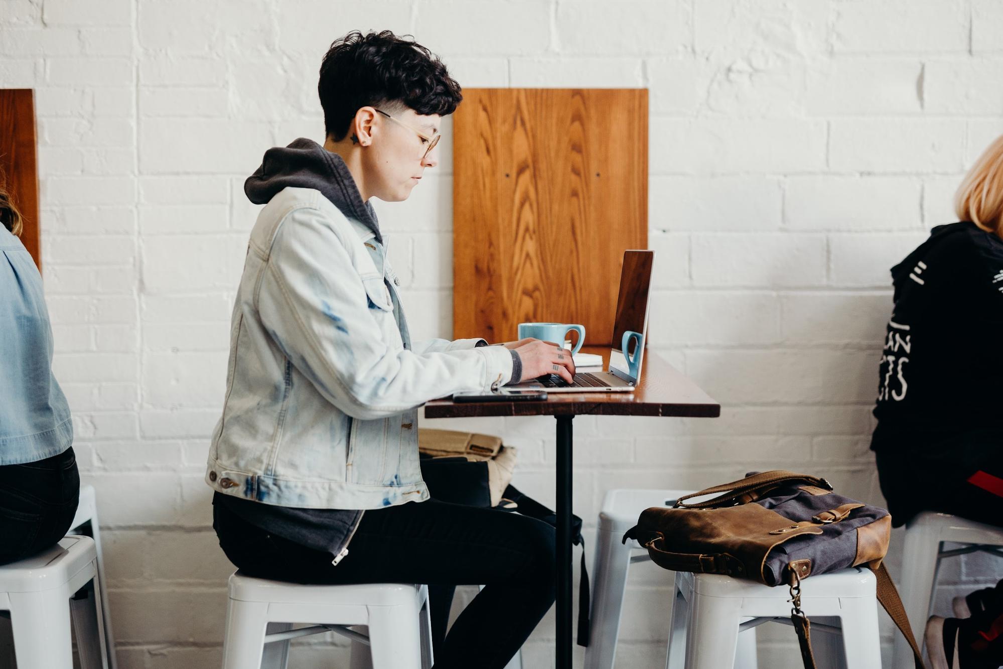 Person wearing a denim jacket sat on a stool in a coffee shop working on a laptop.