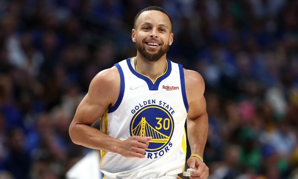Steph Curry of the Golden State Warriors smiling and wearing a jersey on court that has a prominent Rakuten logo