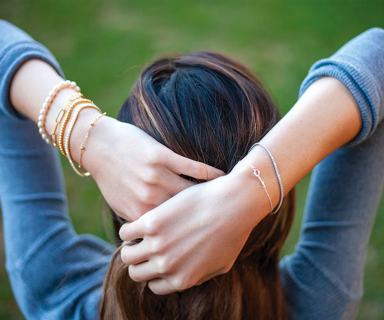 Woman in field, her hands on hair, wrists wearing thin bracelets of silver, gold, and pearl chains.