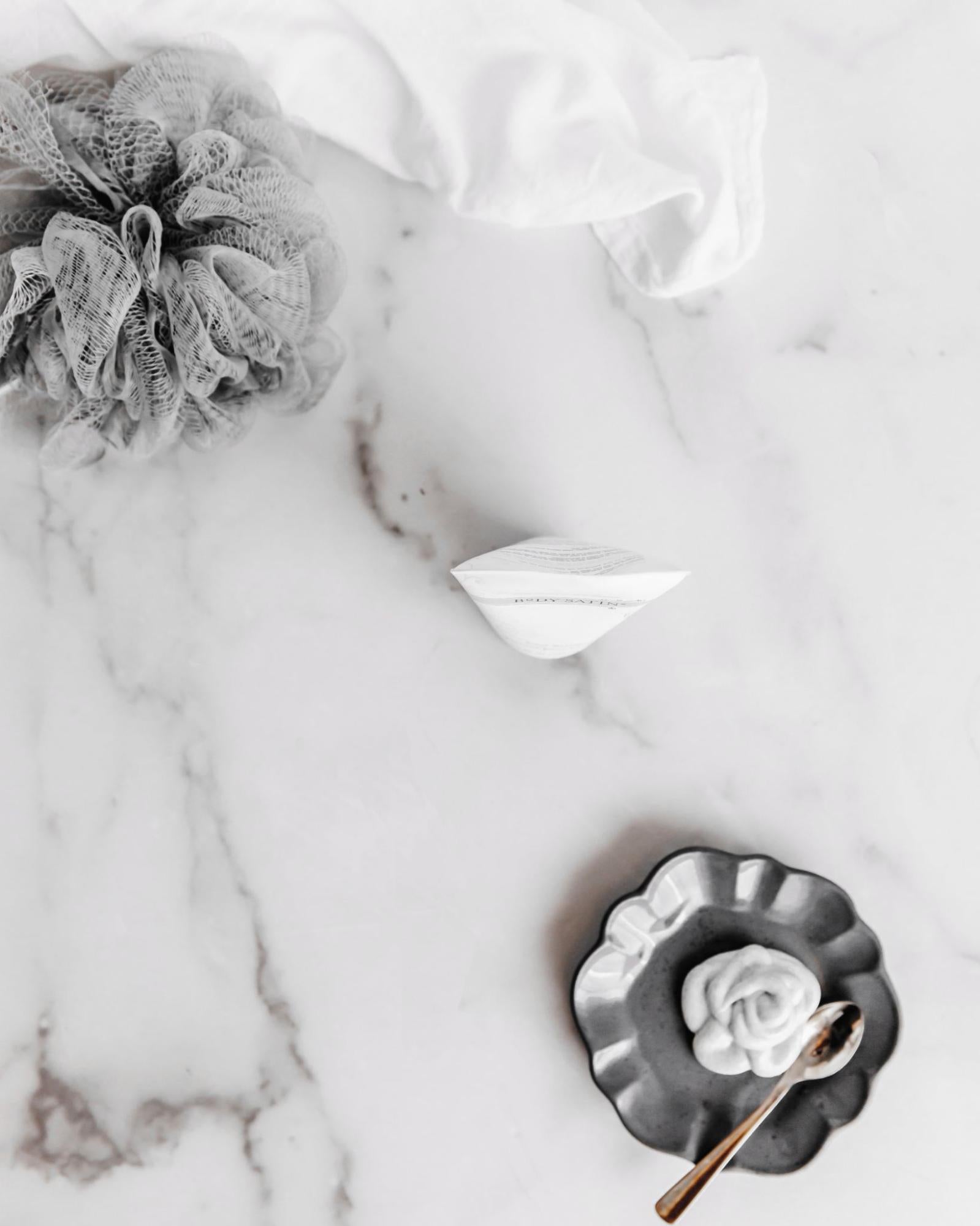 Same overhead shot of luffa, towel, and bottle, but now there is a silver plate with a dollop of cream in the bottom right corner