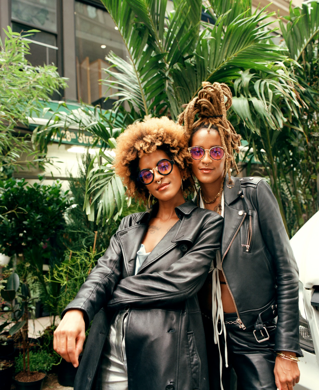 an image of Coco and Breezy posing in leather jackets, backdropped by large palm plants