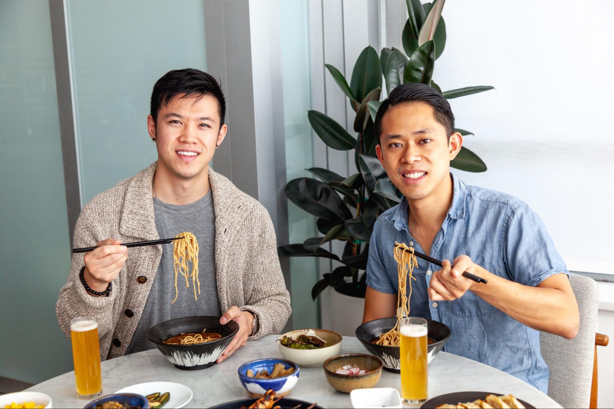 Founders of immi, Kevin Lee and Kevin Chanthasiriphan sit at a dinning table enjoying bowls of various noodles.