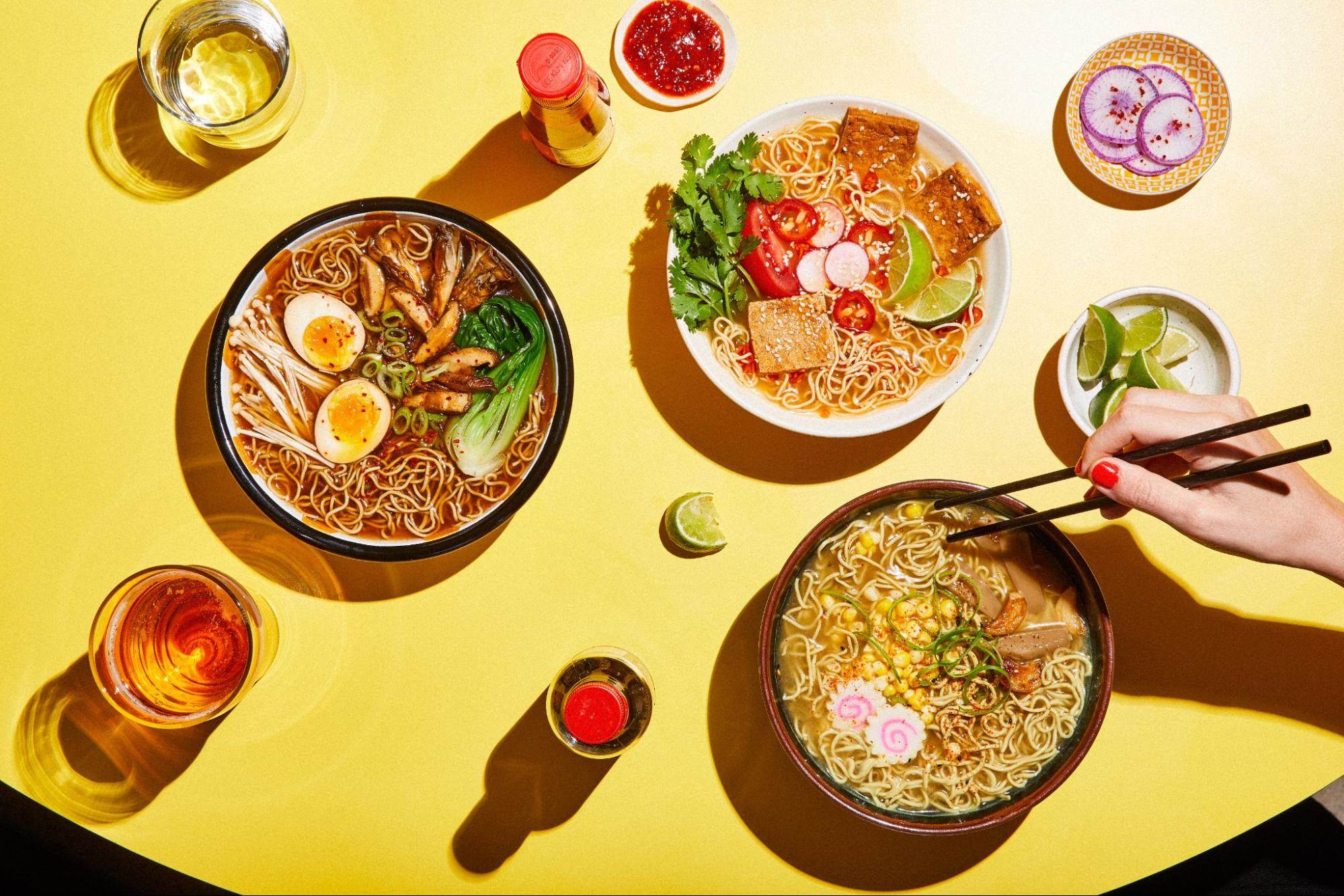 Three different bows of ramen on a yellow table with a hand holding chopsticks in the frame.